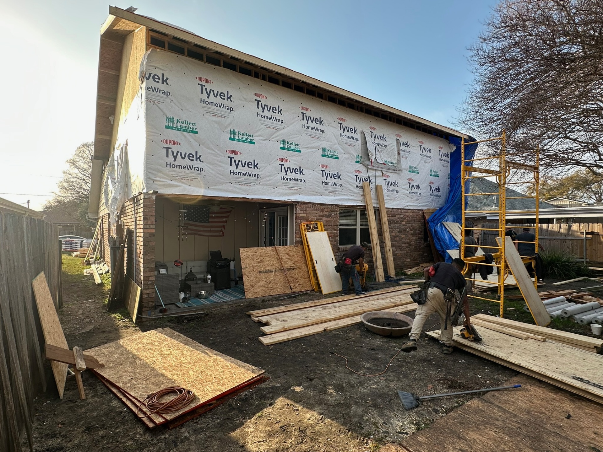 Rear elevation of Metairie home addition mid-wrap with Tyvek installed over second-floor sheathing while crew cuts framing lumber and sets scaffolding