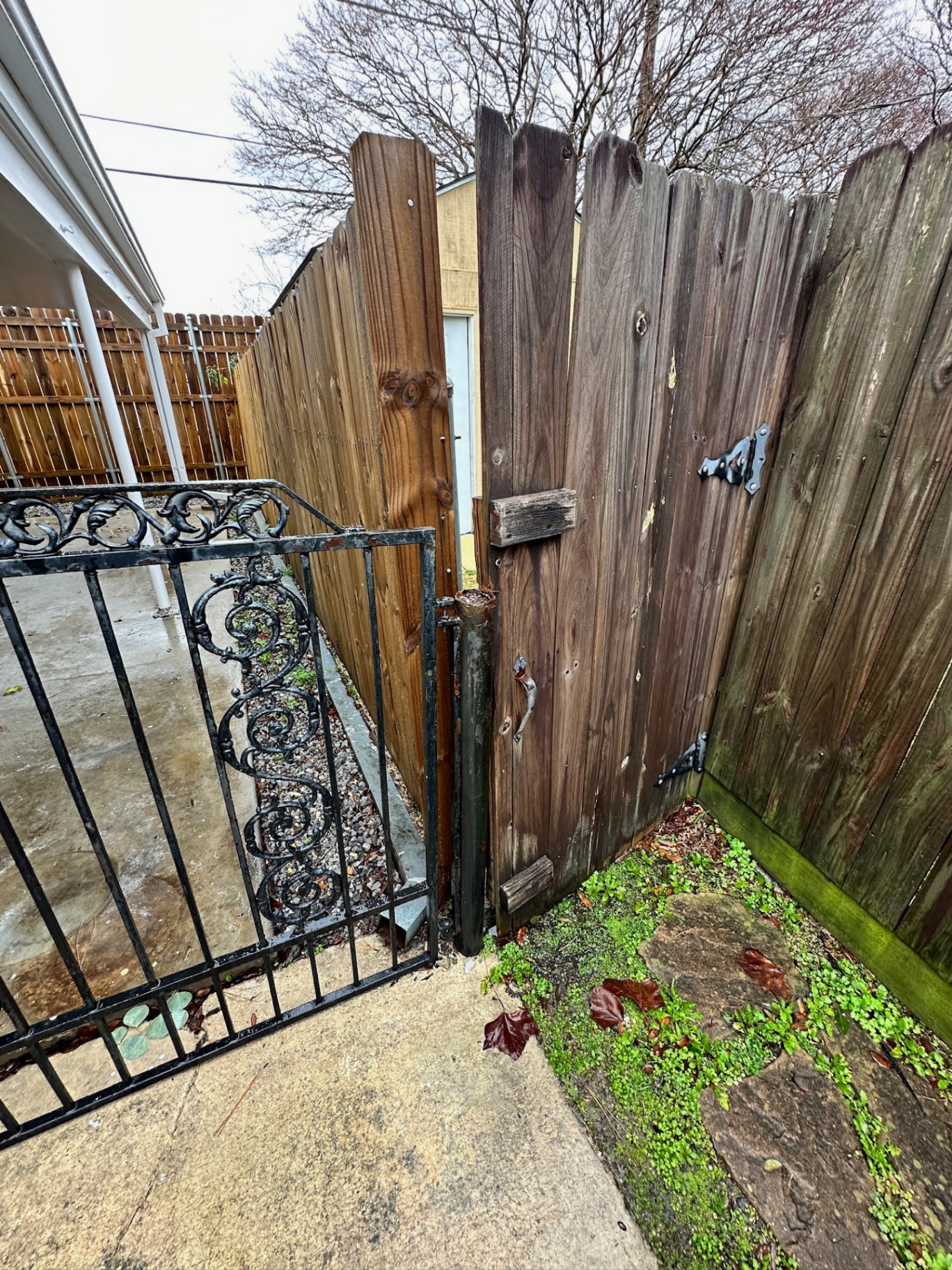 Before photo — original weathered wood gate next to the wrought iron carport gate at the Metairie home, showing rusted hinges, split boards and a leaning post