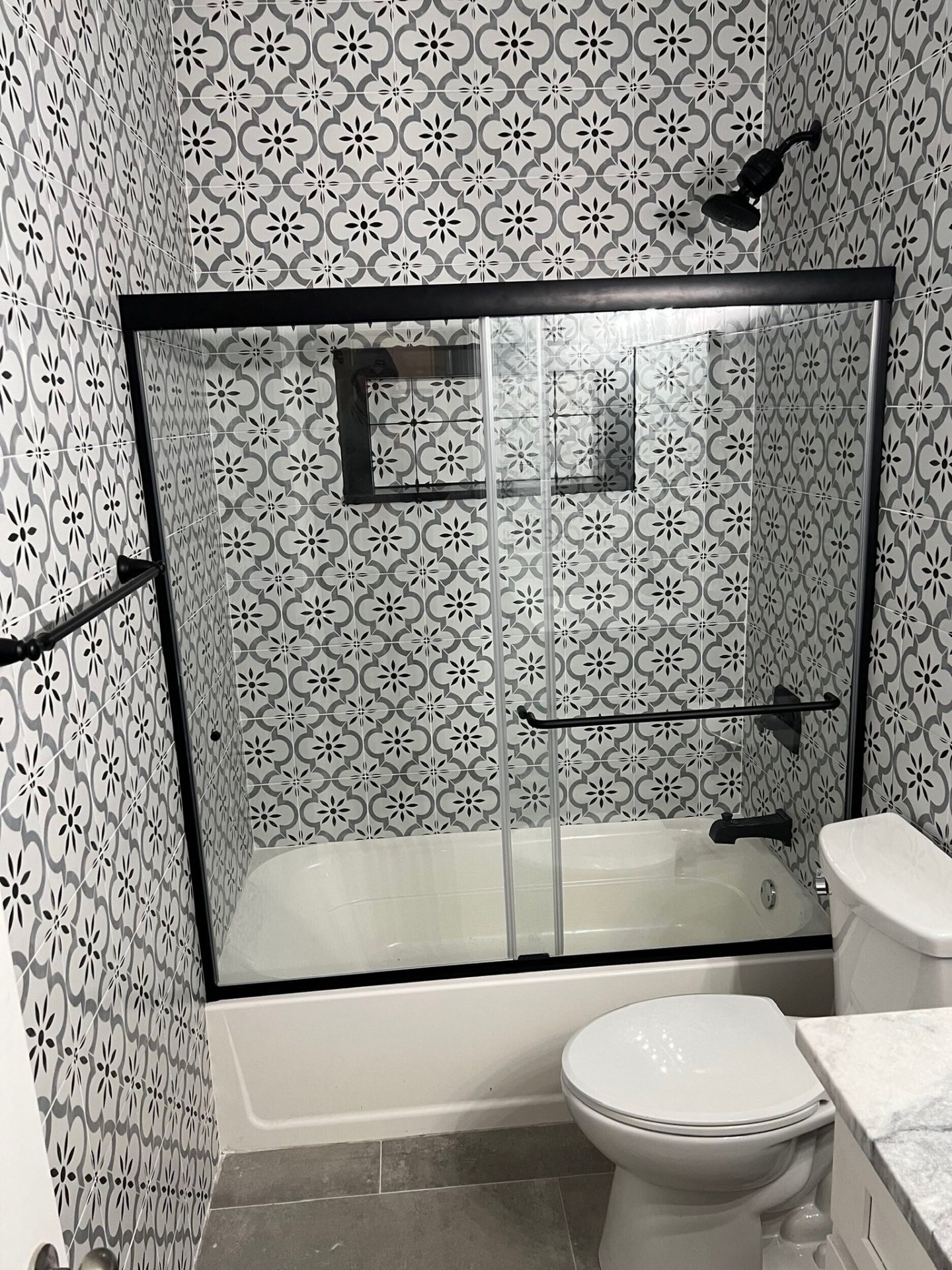 Straight-on view of finished tub and shower surround in Meraux LA bathroom renovation with patterned porcelain tile from floor to ceiling, recessed niche, matte black shower head and tub filler, and framed sliding glass tub door
