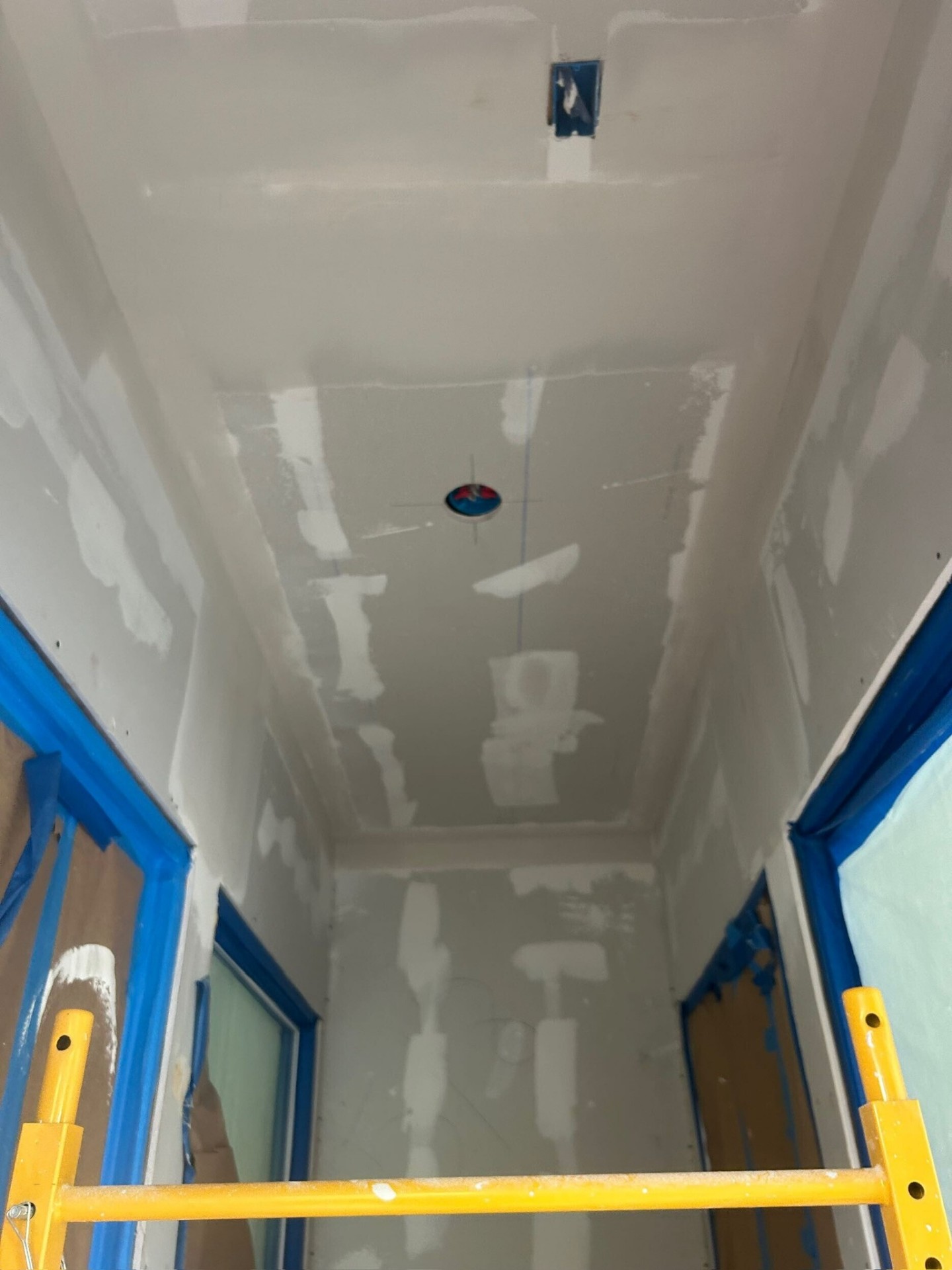 Looking up into the new vaulted stairwell of the Metairie second-floor addition with drywall hung and seams taped on the tall walls and sloped ceiling, recessed can light rough-in centered overhead and yellow scaffolding staged below