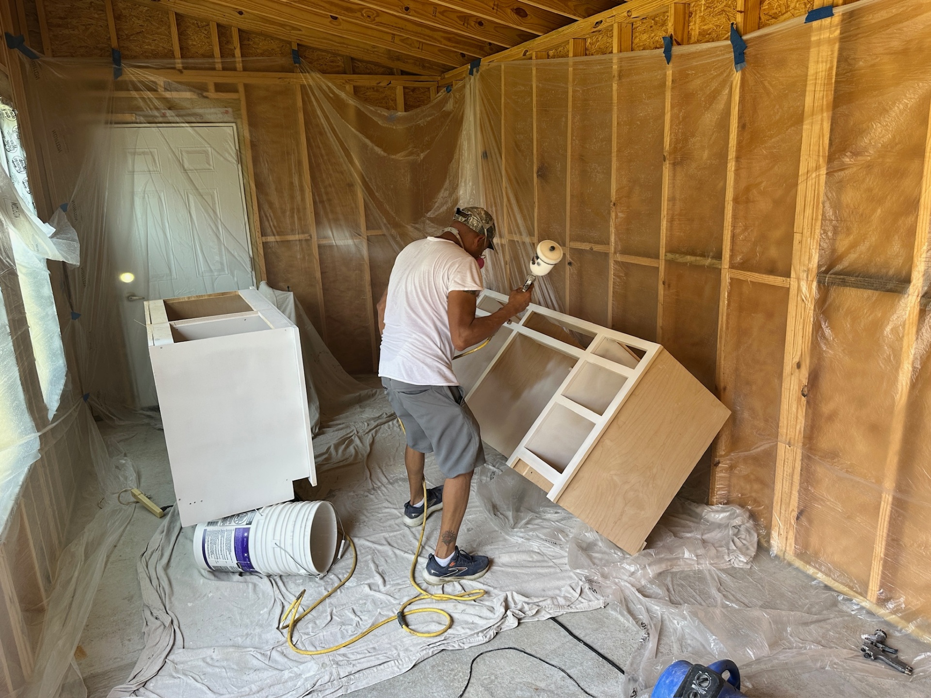 Finisher spraying the side of a custom vanity cabinet with HVLP gun inside a plastic-draped spray booth at the Metairie master bath project