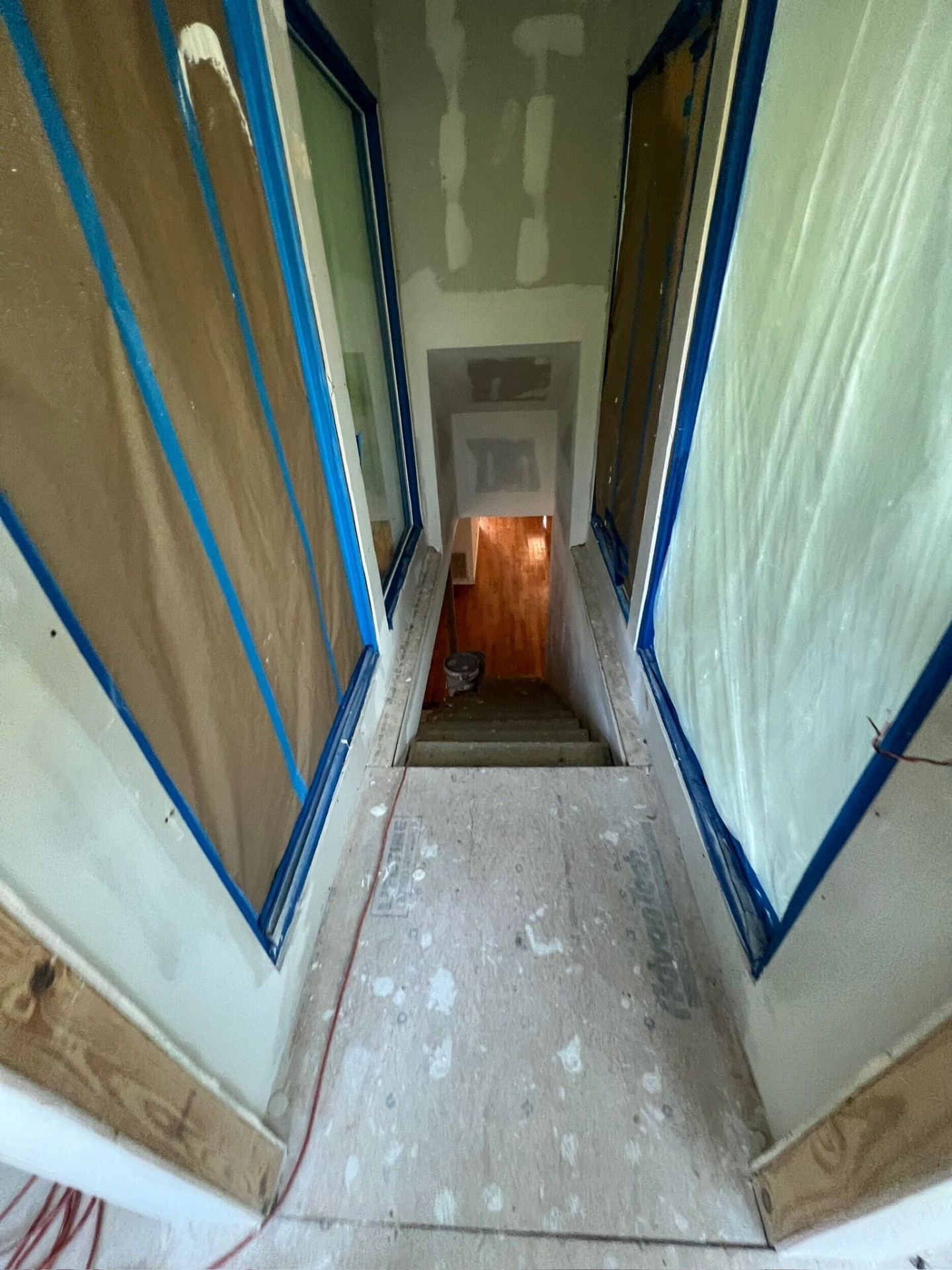 Looking down the new second-floor stairwell of the Metairie addition with blue painters tape masking the trim, finished drywall walls and the original hardwood floor visible at the bottom landing