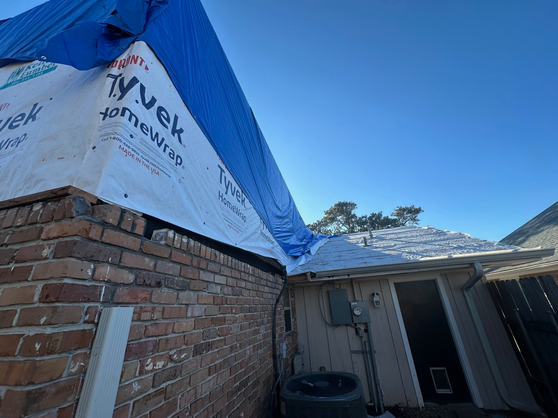 Exterior corner view of Metairie home where new Tyvek-wrapped second-floor sheathing meets original brick first floor with blue tarp draped over the roof transition and Kellett Lumber Tyvek branding visible