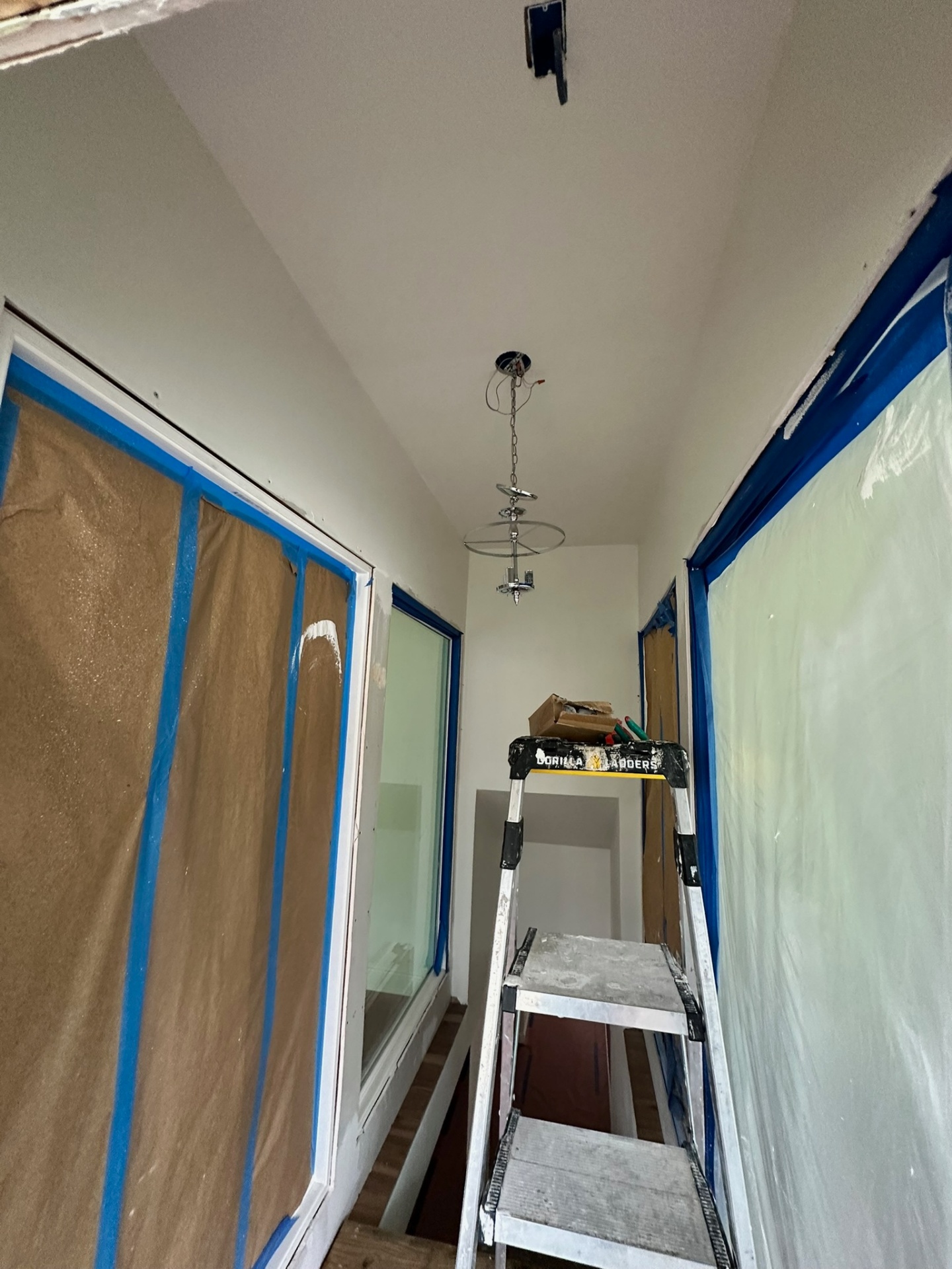 New vaulted stairwell on the Metairie second-floor addition fully masked with brown ram board and blue tape over the side windows, crystal pendant chandelier hanging from the ceiling and a Gorilla ladder staged for ceiling paint cut-in