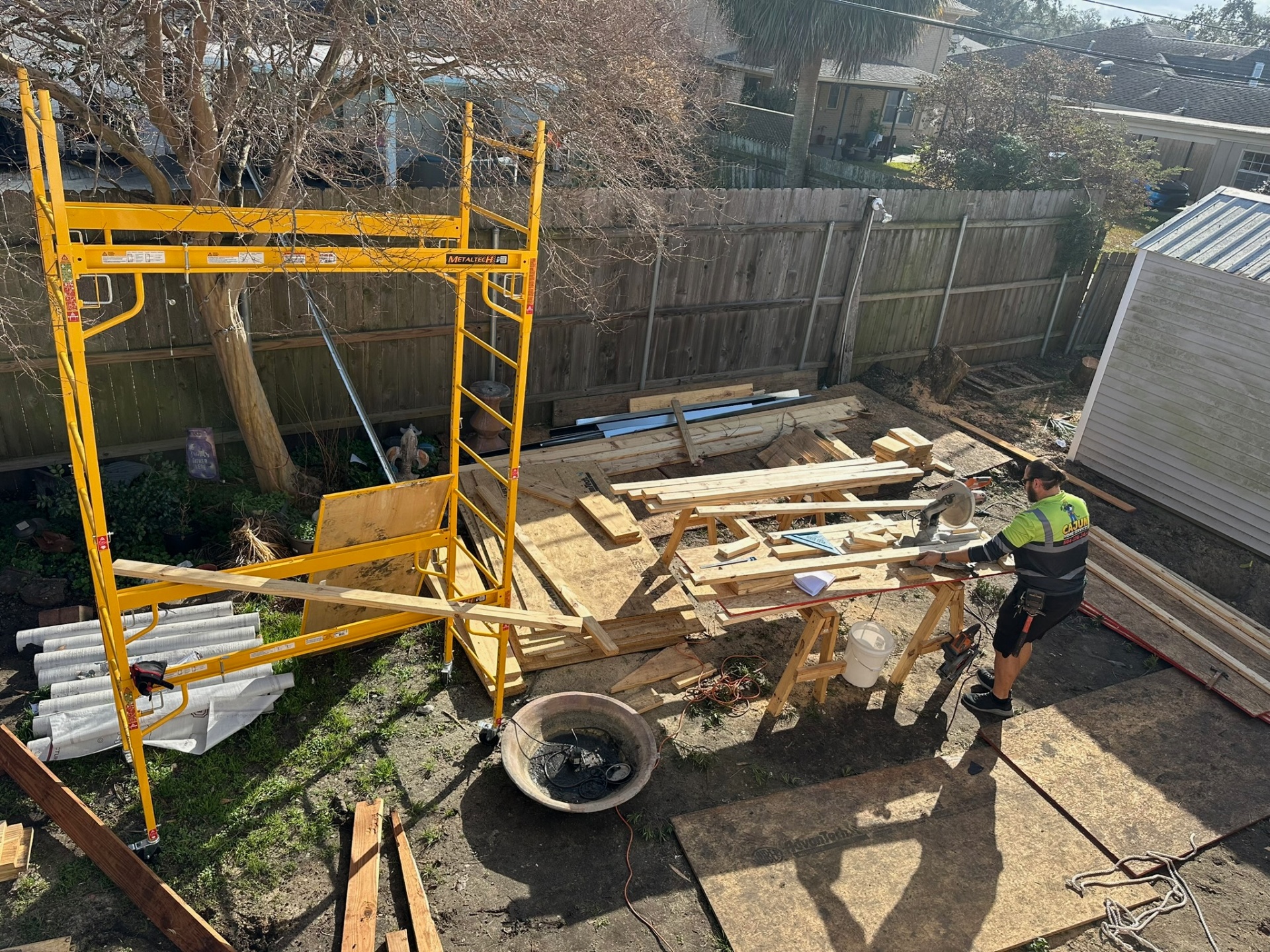 Aerial backyard view of Metairie second-floor addition site with Cajun Home Services carpenter in branded shirt cutting framing lumber on sawhorses next to a scaffold tower, OSB sheets and rolls of Tyvek staged for the build