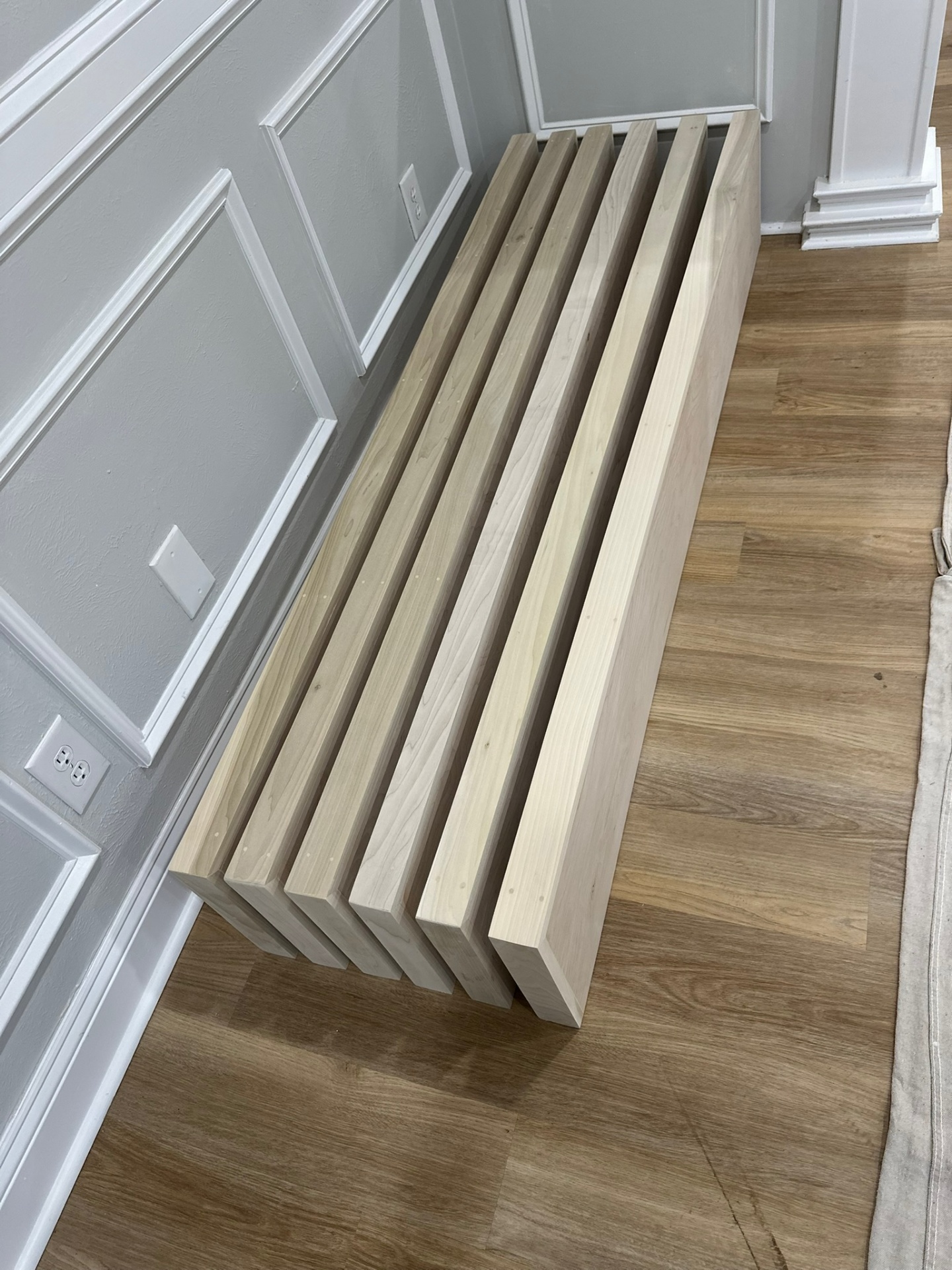 Six raw maple floating shelves laid flat and staggered on the LVP floor against gray wainscot paneling in a Meraux Louisiana living room before staining