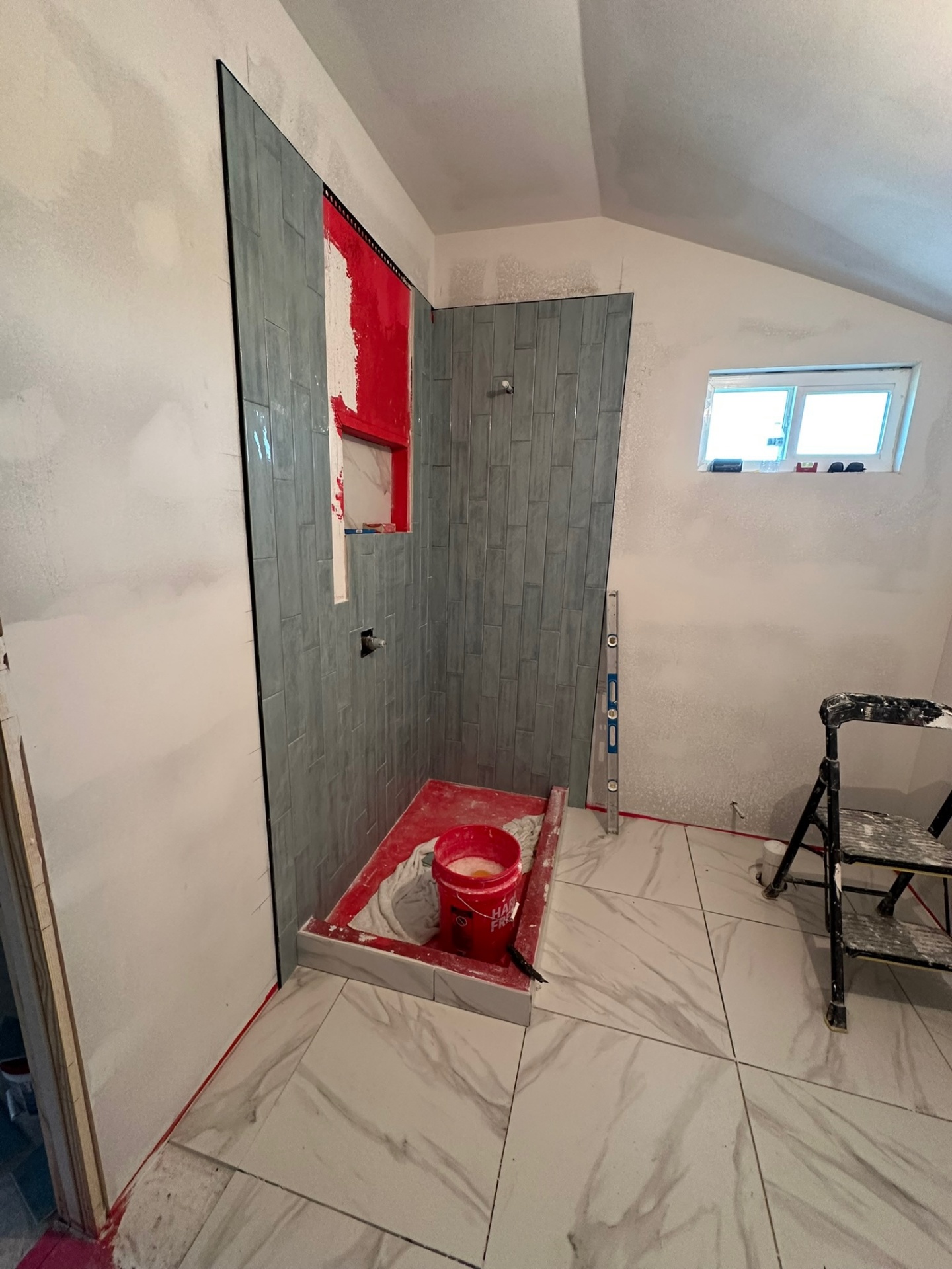 Wide view of the new master bathroom in the Metairie second-floor addition during tile work showing the vaulted gable ceiling, sage-green vertical shower tile, marble-look floor tile, a vent window and ladder staged for the tile crew