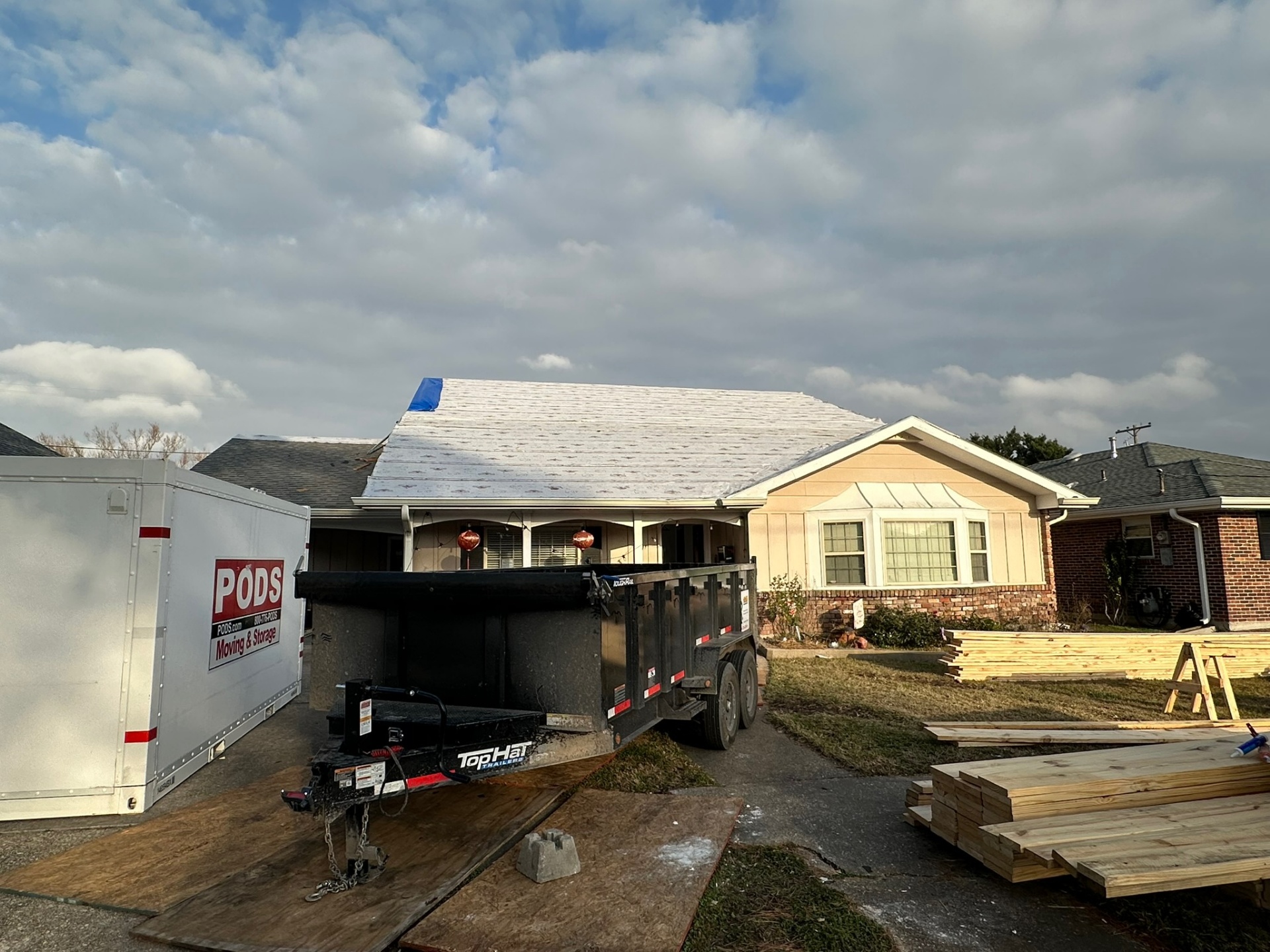 Front elevation of original single-story Metairie home with synthetic underlayment over existing roof, PODS container, dump trailer and lumber staged for second-floor addition