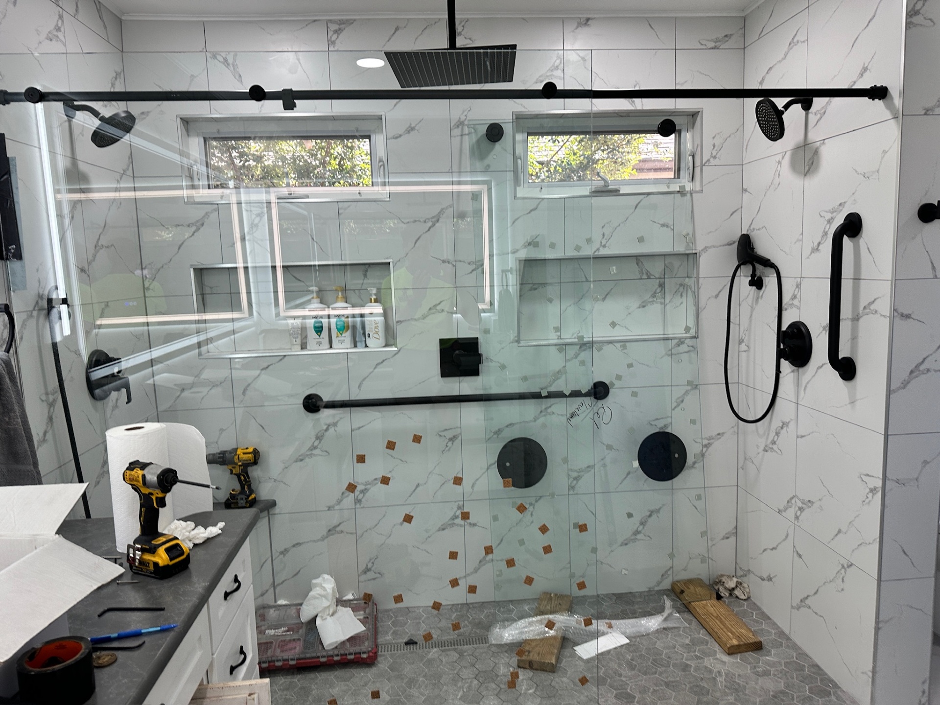Frameless glass shower doors installed with protective decals, black rain head, dual shower heads, and grab bars in Metairie master bath