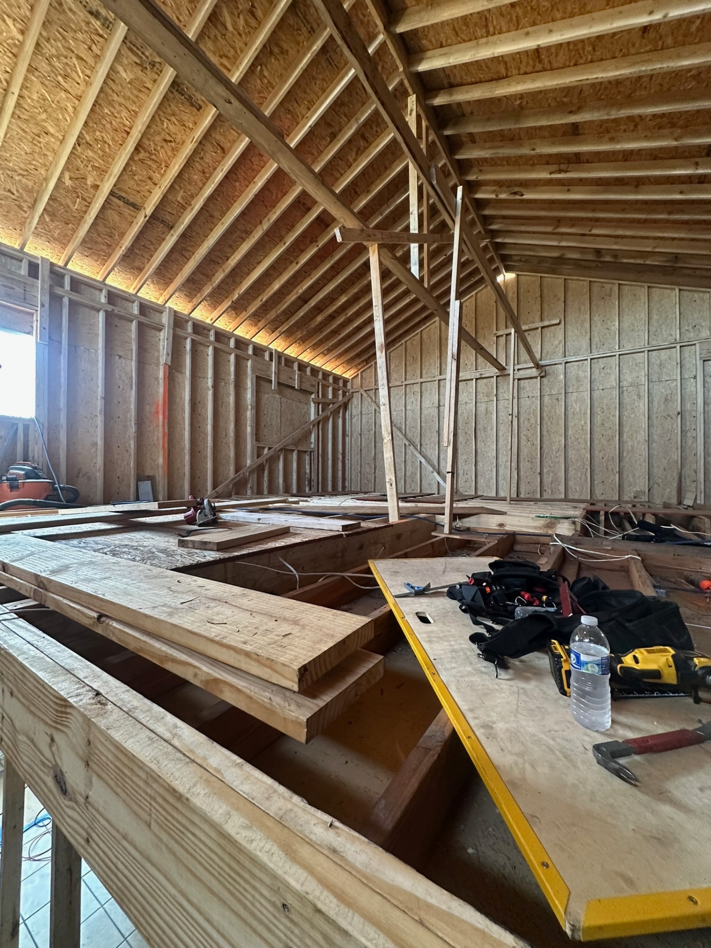 Long interior view down Metairie second-floor addition with new roof rafters overhead, OSB sheathed gable wall at the far end and tools staged on temporary walking boards