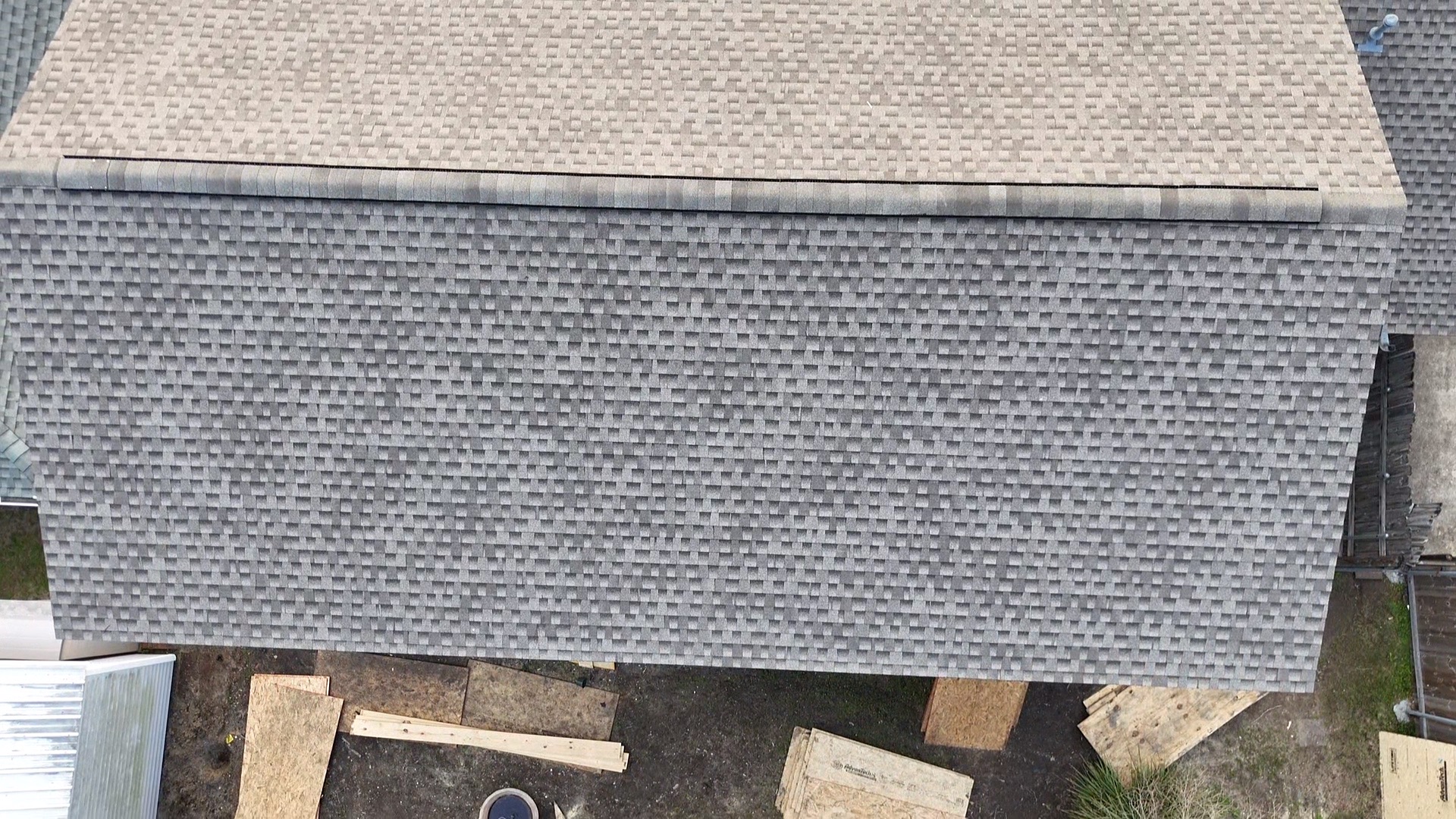 Close aerial detail of the ridge transition between new tan architectural shingles on the second-floor addition and existing gray shingles on the original single-story roof below