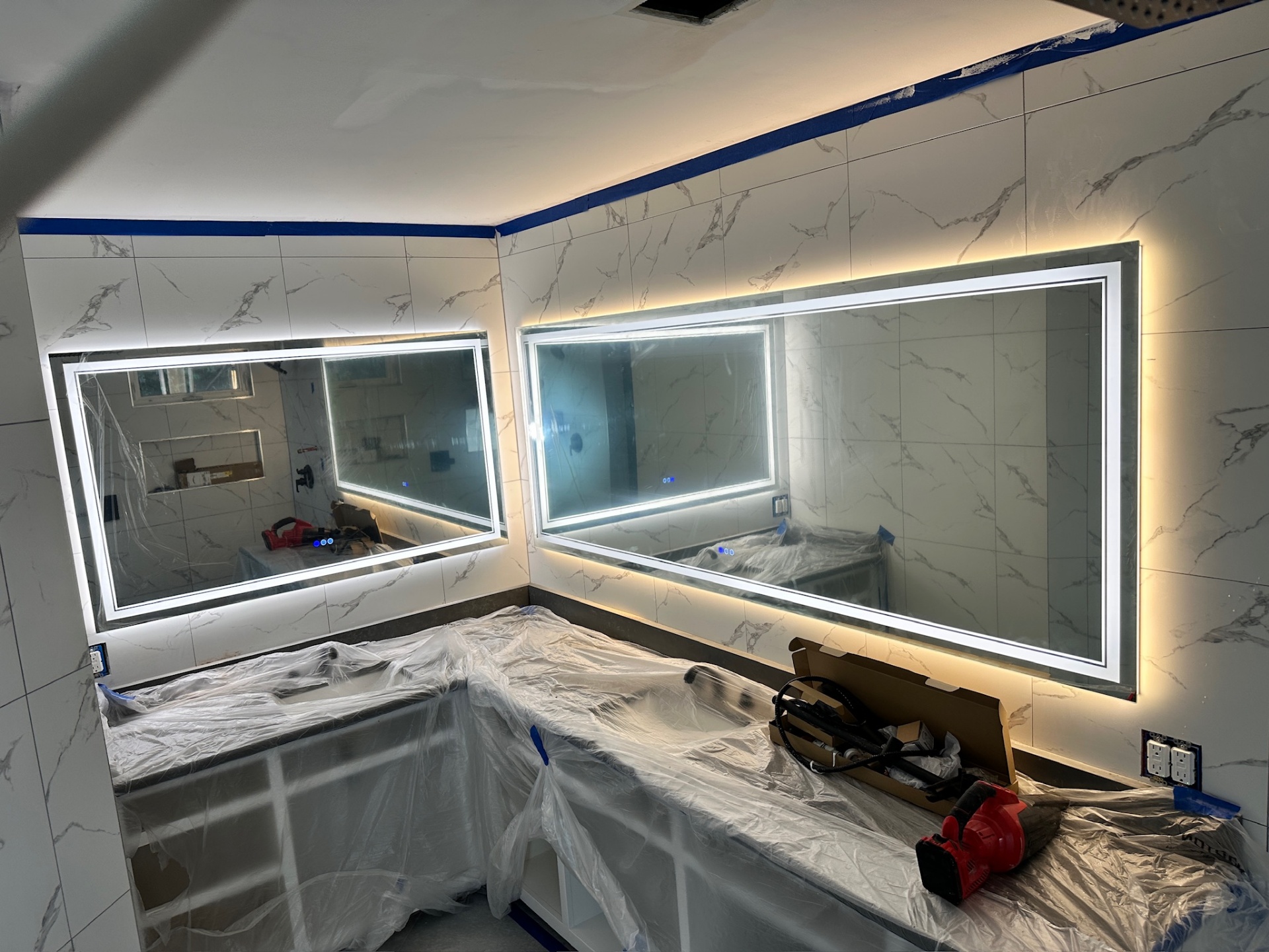 Dual LED-backlit vanity mirrors mounted and lit against marble-look tile with counters masked for paint in Metairie master bath