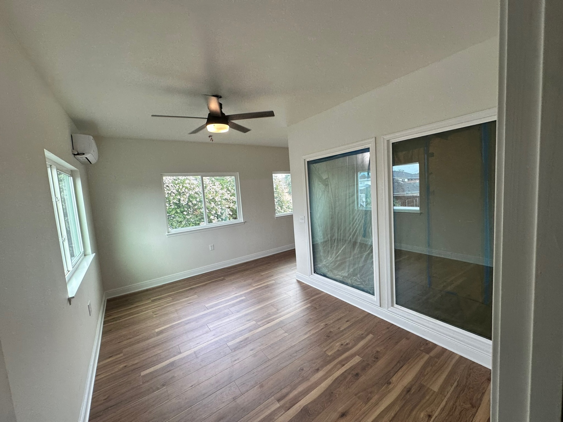 Finished second-floor loft / living area on the Metairie addition with wide-plank LVP flooring, ceiling fan, ductless mini-split, side window, large rear sliding glass doors masked with plastic during punch-out and view into adjoining finished room