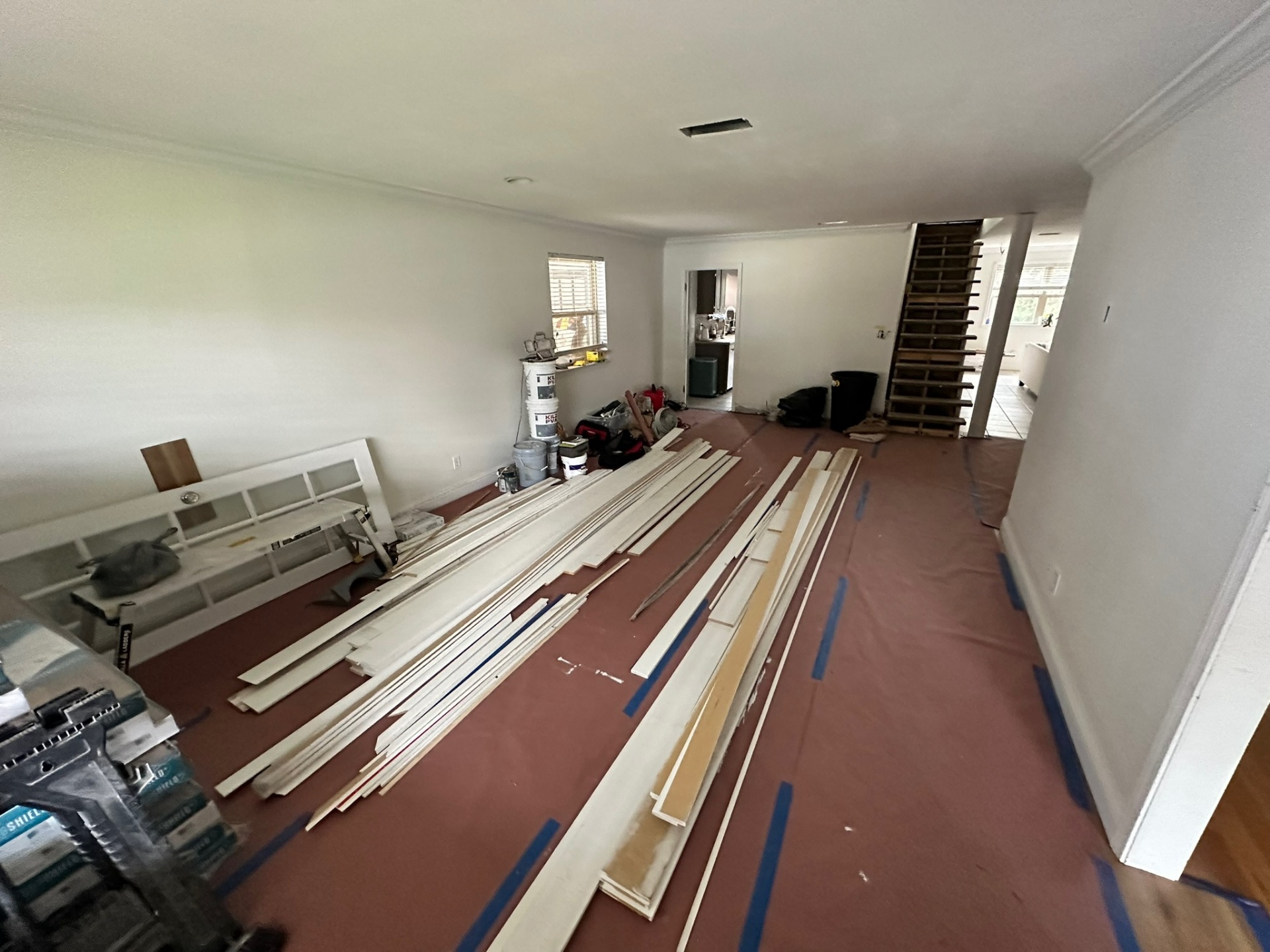 First-floor living area of the Metairie home during the second-floor addition with red rosin paper floor protection, blue tape seams and stacks of primed baseboard and trim staged on the floor next to tile saw and tool bags