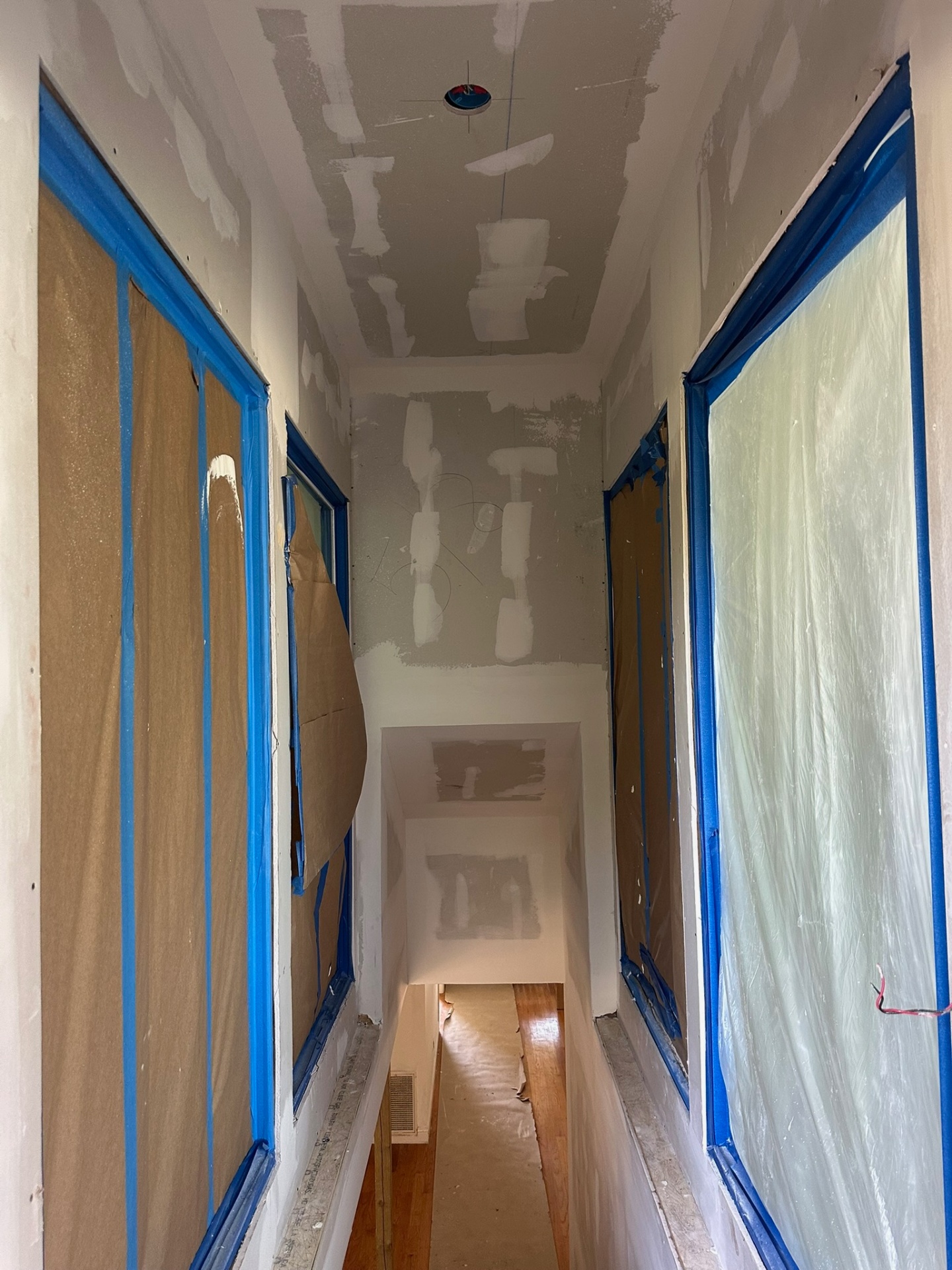 Narrow new second-floor hallway on the Metairie addition with drywall hung, ceiling seams taped and floated, windows and doors masked with blue painters tape and brown paper, looking down toward the stair landing