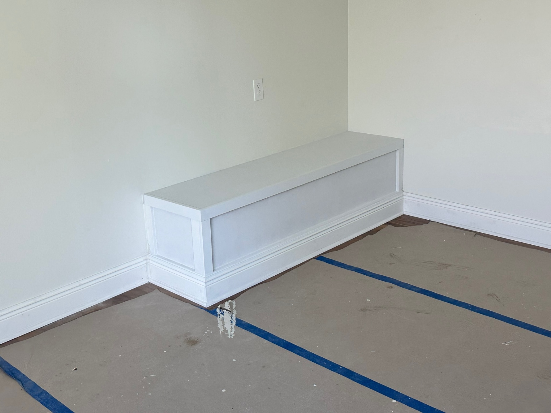 Custom-built finished window bench seat with shaker panel front, painted white, base shoe and baseboard trim, installed in second-floor bedroom over engineered wood flooring