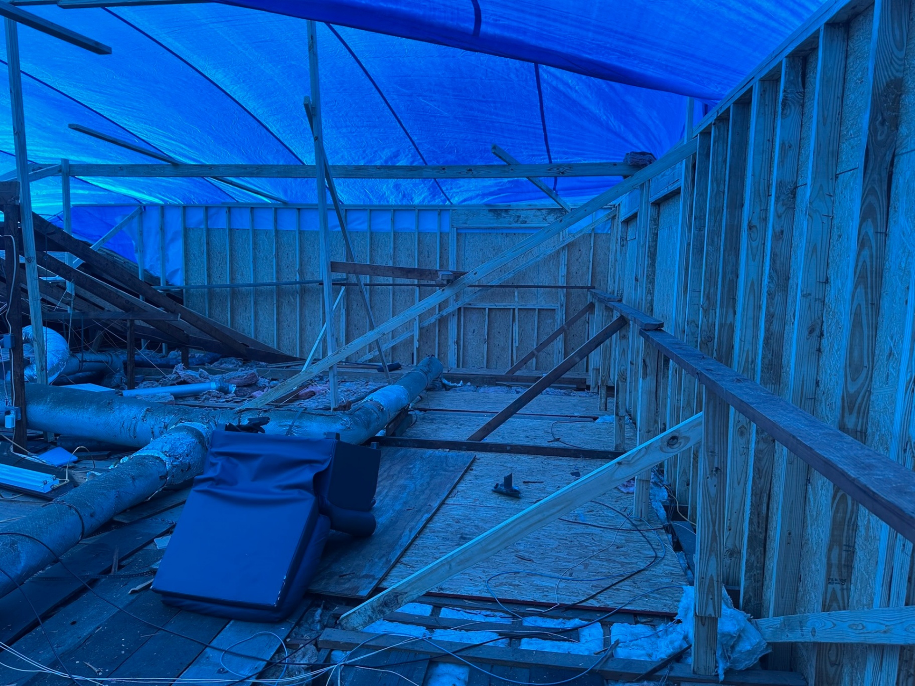Inside Metairie attic during second-floor tear-off with blue tarp roof overhead, exposed ductwork running across the floor and new stud walls already framed at the perimeter