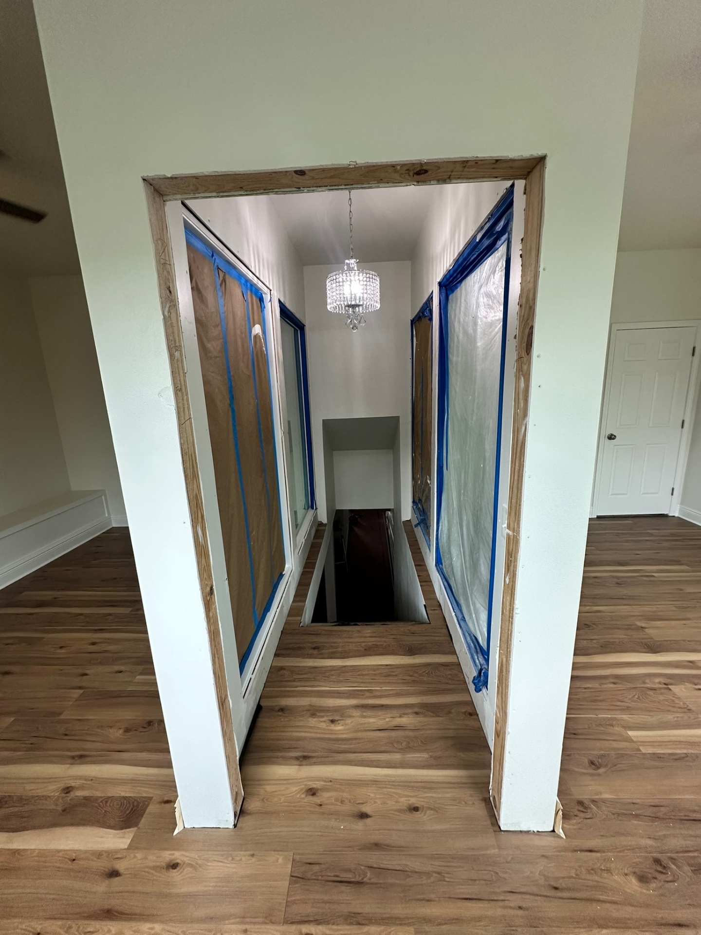 Top of the new stairwell on the second floor of the Metairie addition with raw lumber header still exposed, side walls masked in paper and plastic for paint, crystal drum chandelier hanging over the opening and finished wood-look LVP flooring on the new second floor