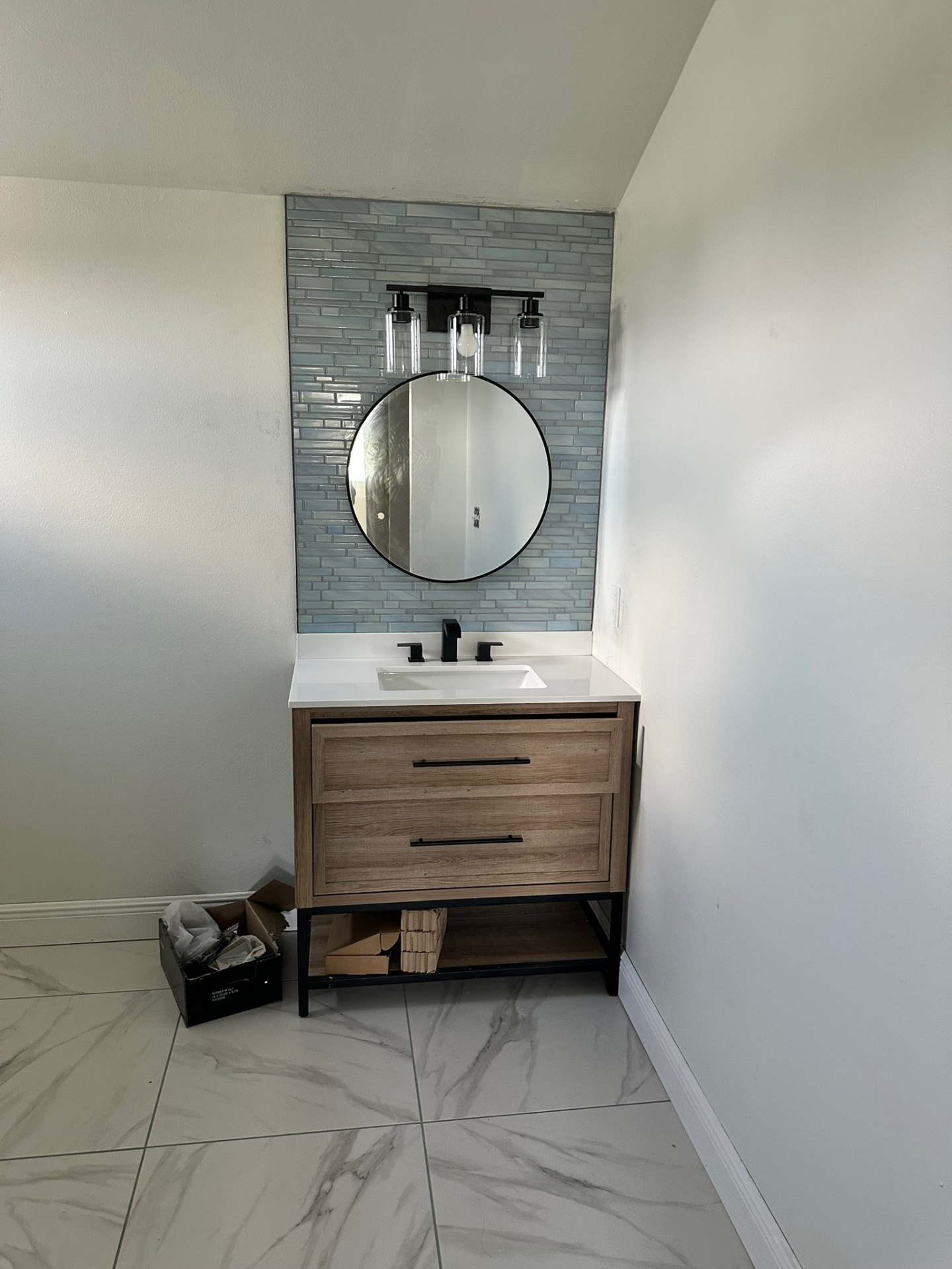 Bathroom vanity with round mirror, glass tile backsplash, and marble-look flooring installed by Cajun Home Services in Metairie LA