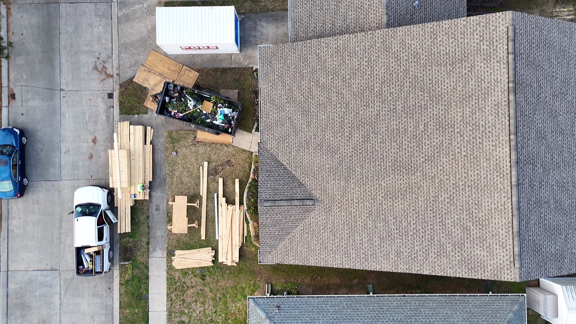 Aerial drone view of original single-story Metairie home with shingle roof, PODS container in driveway and lumber staged in yard before second-floor addition begins
