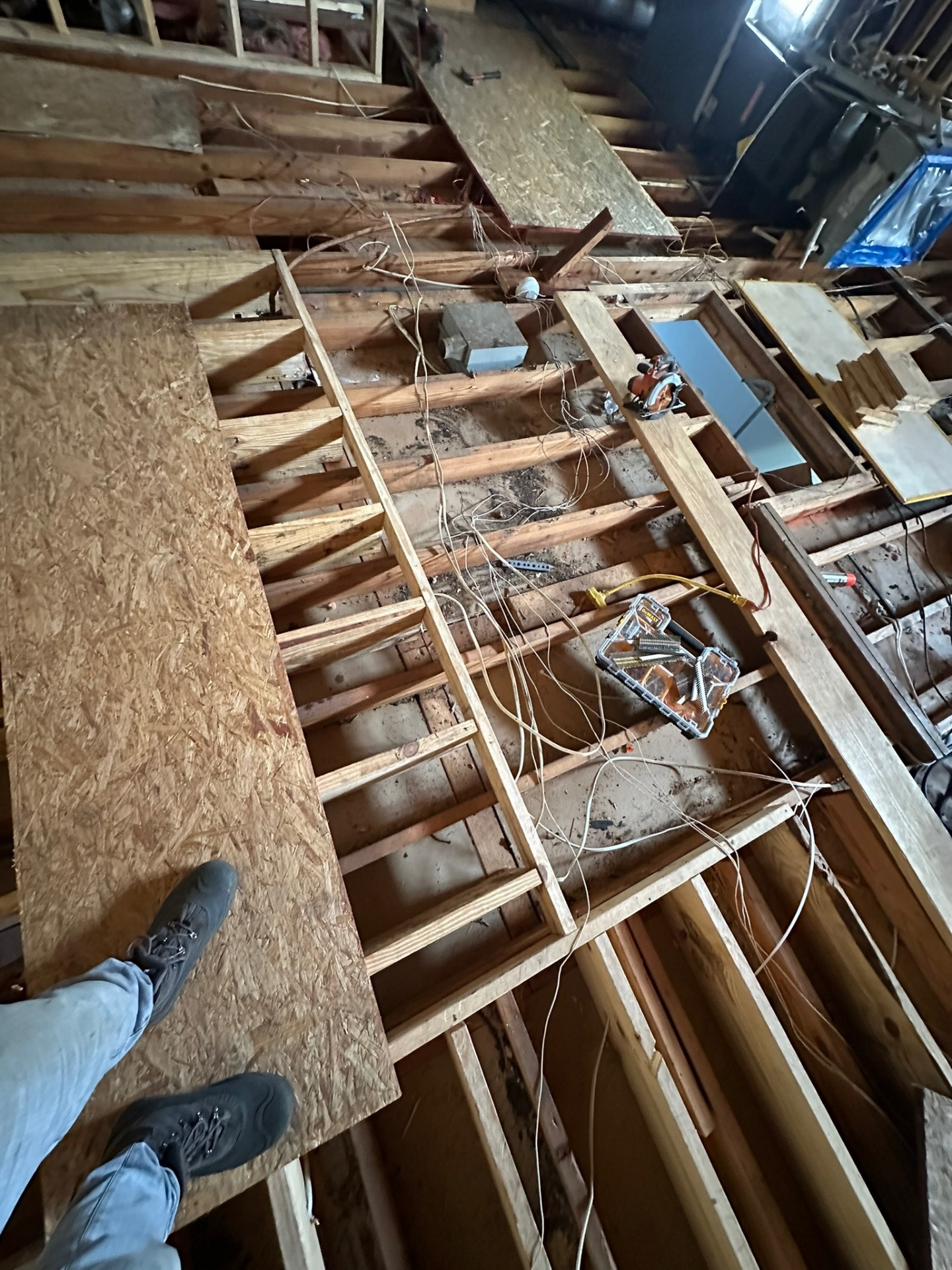 First-person overhead view from inside Metairie attic during demo showing original ceiling joists exposed, old wiring pulled out and worker boots on temporary OSB walking board