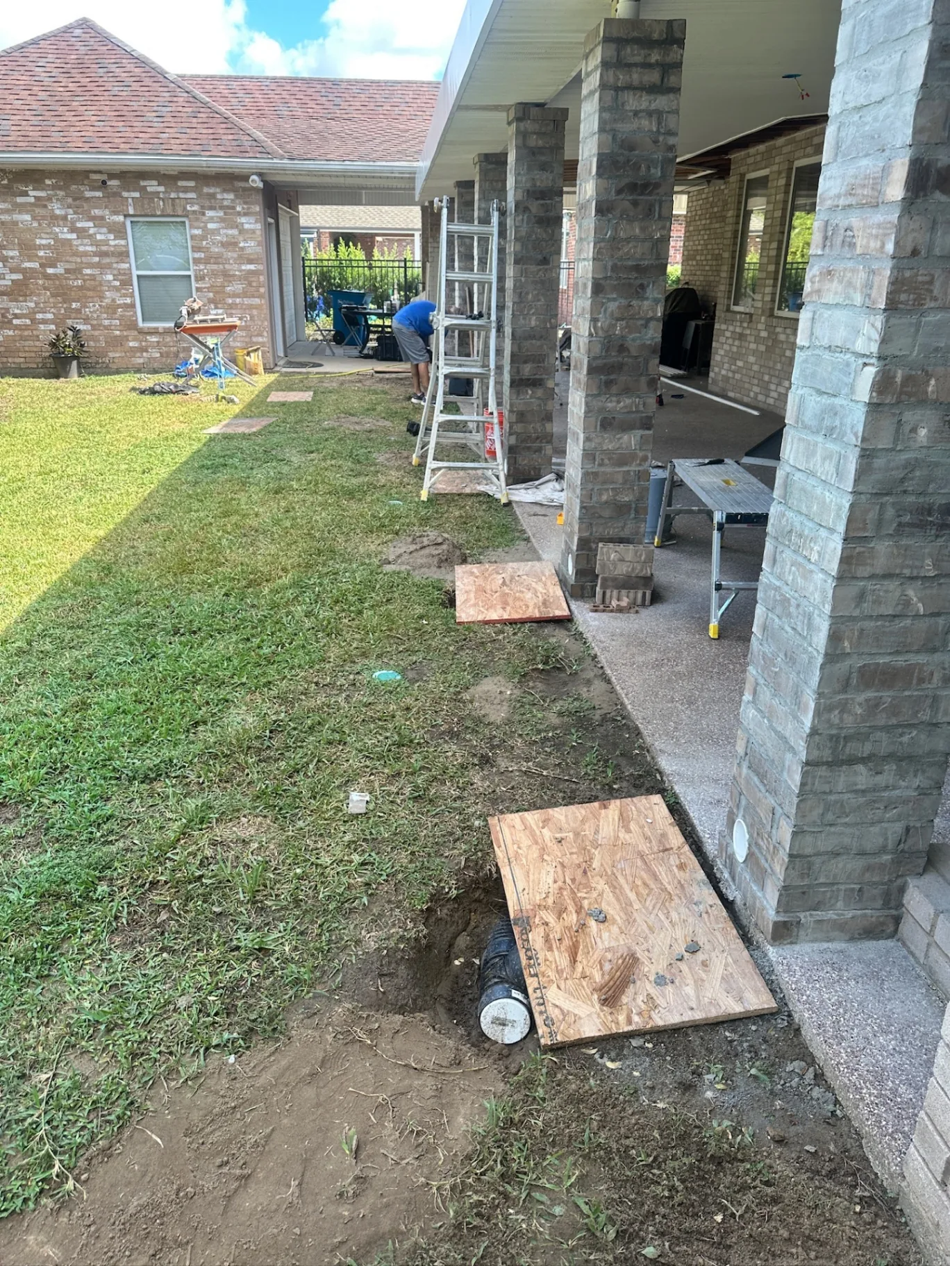Side yard view of newly brick-wrapped patio posts with drain piping exposed for hidden gutter and downspout work at Destrehan Louisiana home