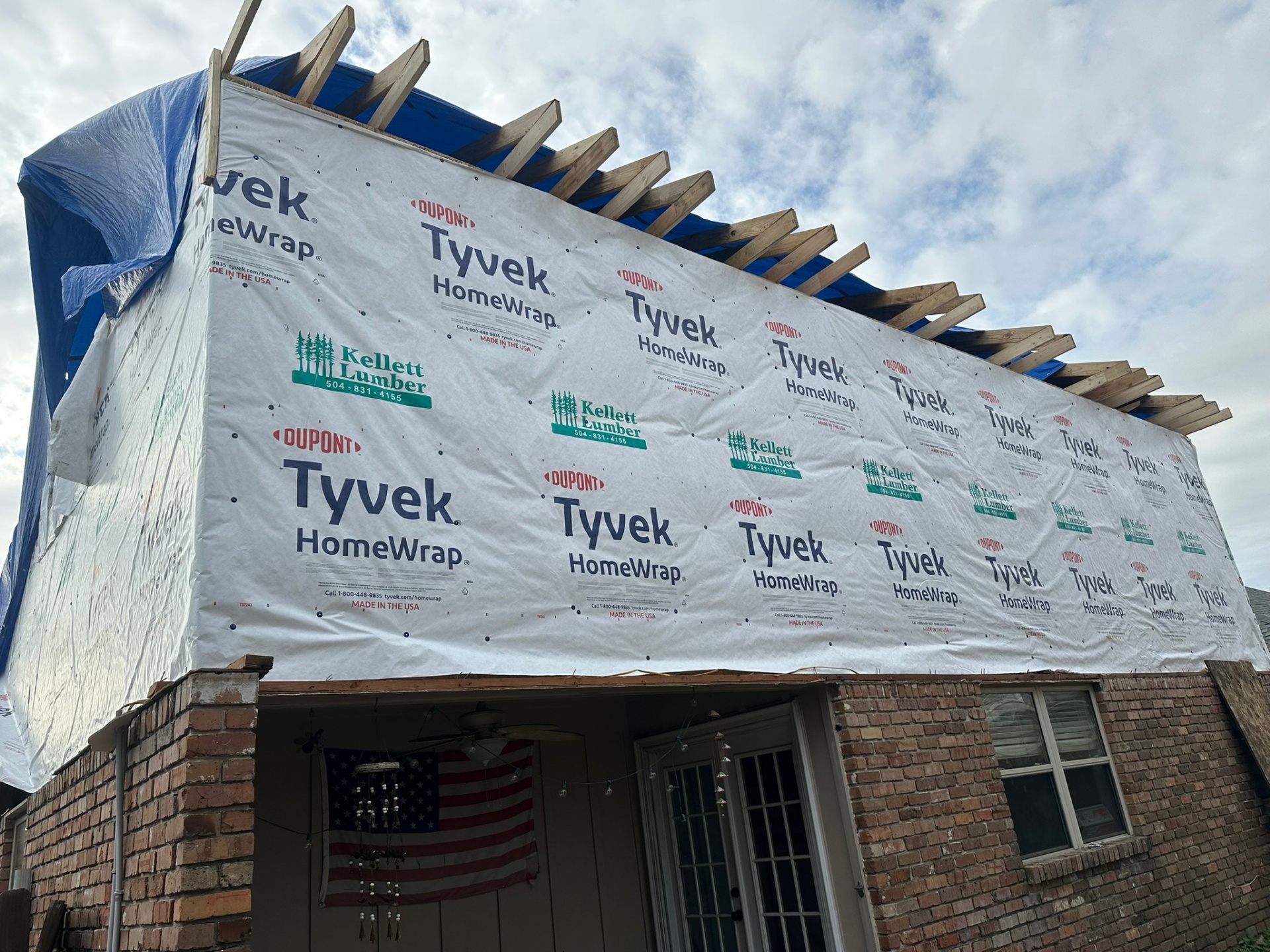 Rear elevation of Metairie second-floor addition with Tyvek wrap installed, exposed rafter tails along the eave and American flag hanging on first-floor porch