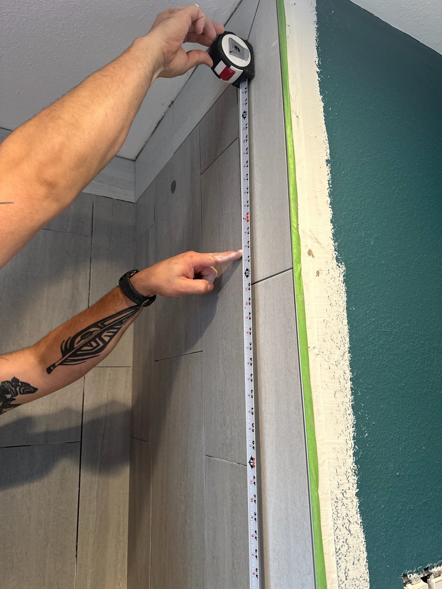 Close-up during shower layout in the Metairie second-floor master bath with a tape measure held against the vertical gray tile field to confirm the cut and reveal at the finished edge before trim-out