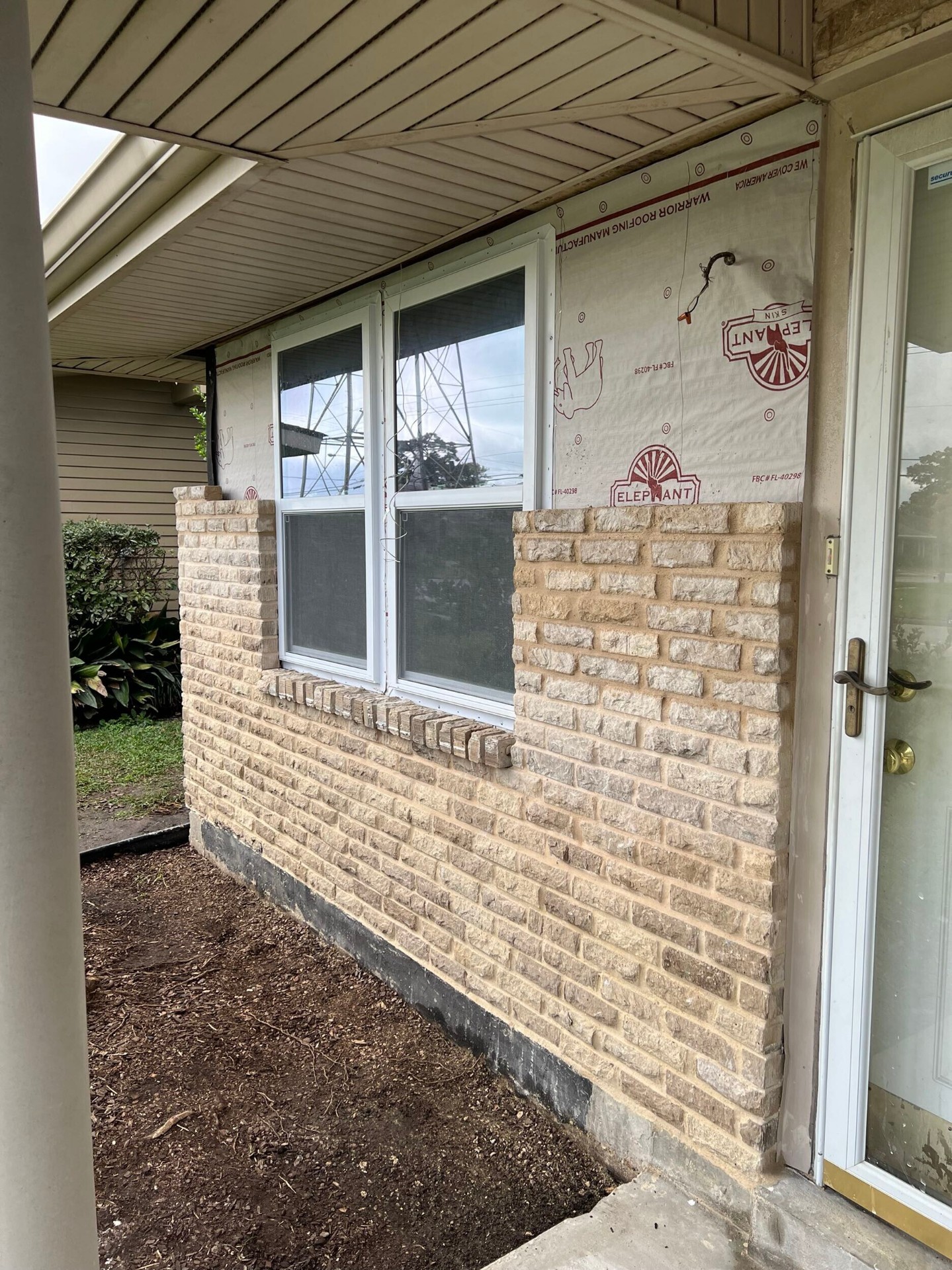Exterior side view of the rebuilt Gretna wall with new brick veneer around a fresh vinyl window and house wrap remaining above for soffit tie-in