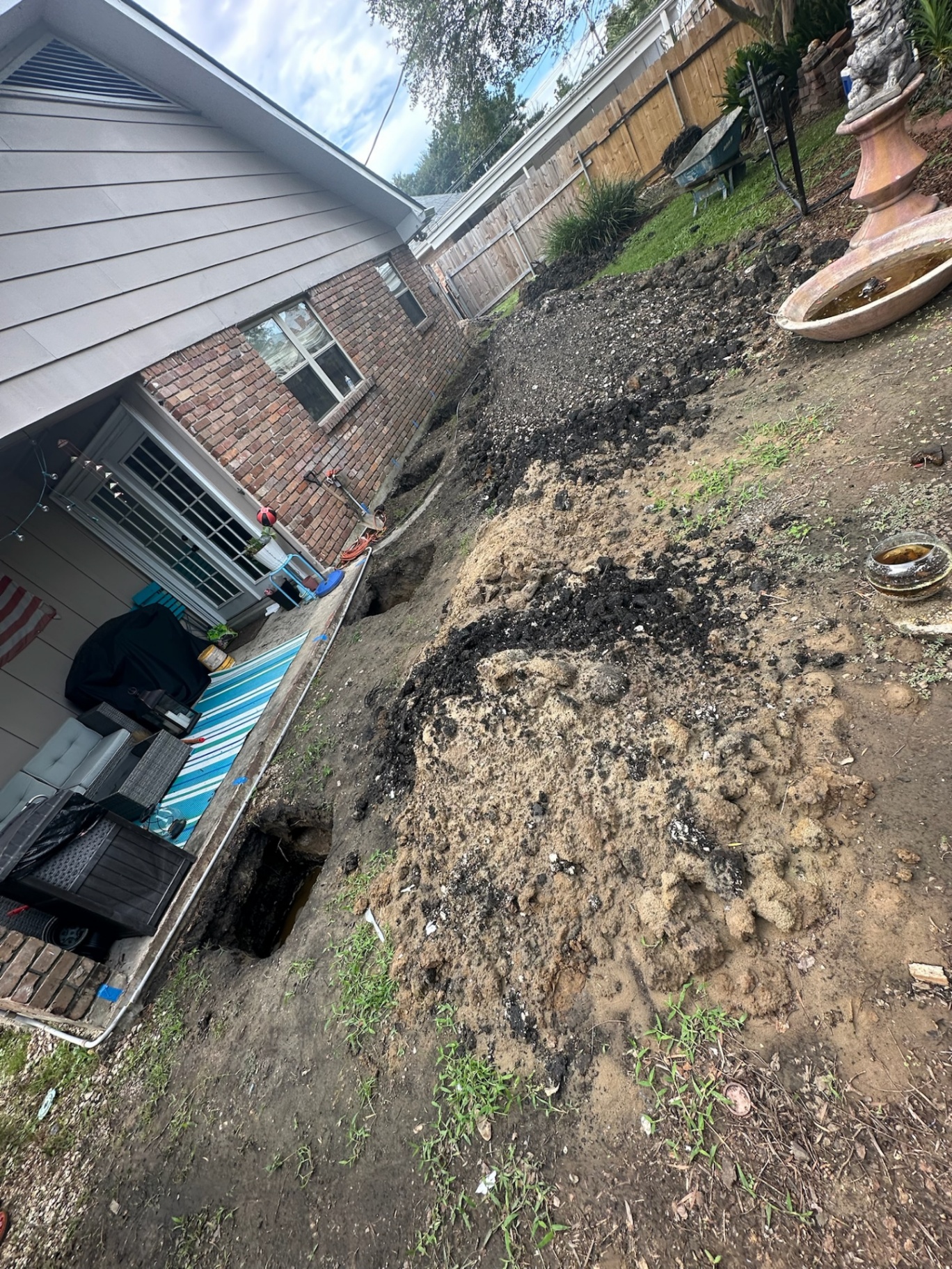 Wide view down the side yard of the Metairie home showing an open excavation trench along the brick foundation with spoil piles and a small open pit at the slab edge during structural foundation prep for the second-floor addition