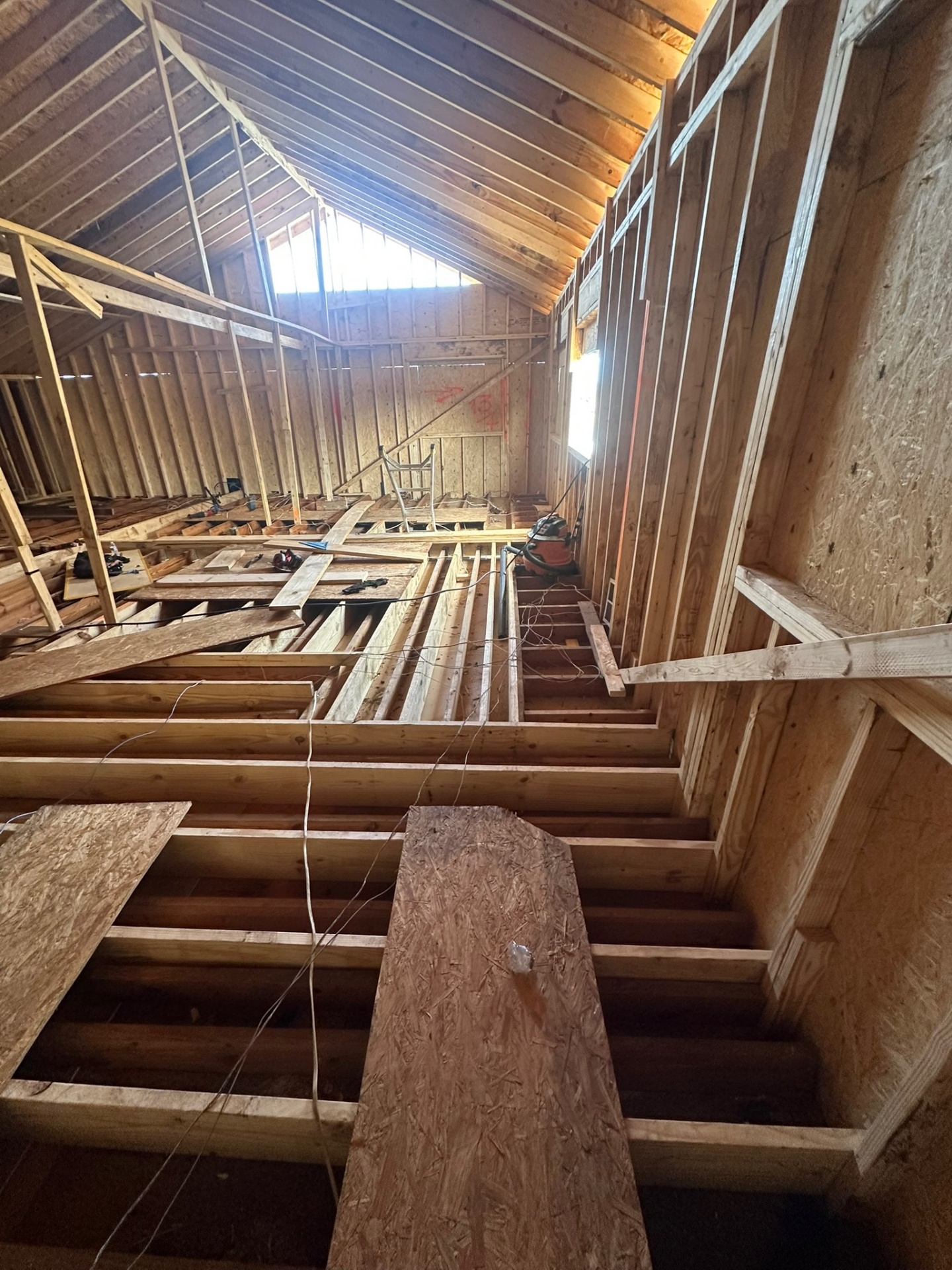 Inside Metairie second-floor framing looking toward sunlit gable end with new roof rafters overhead, knee walls framed along the eaves and partial floor decking in place