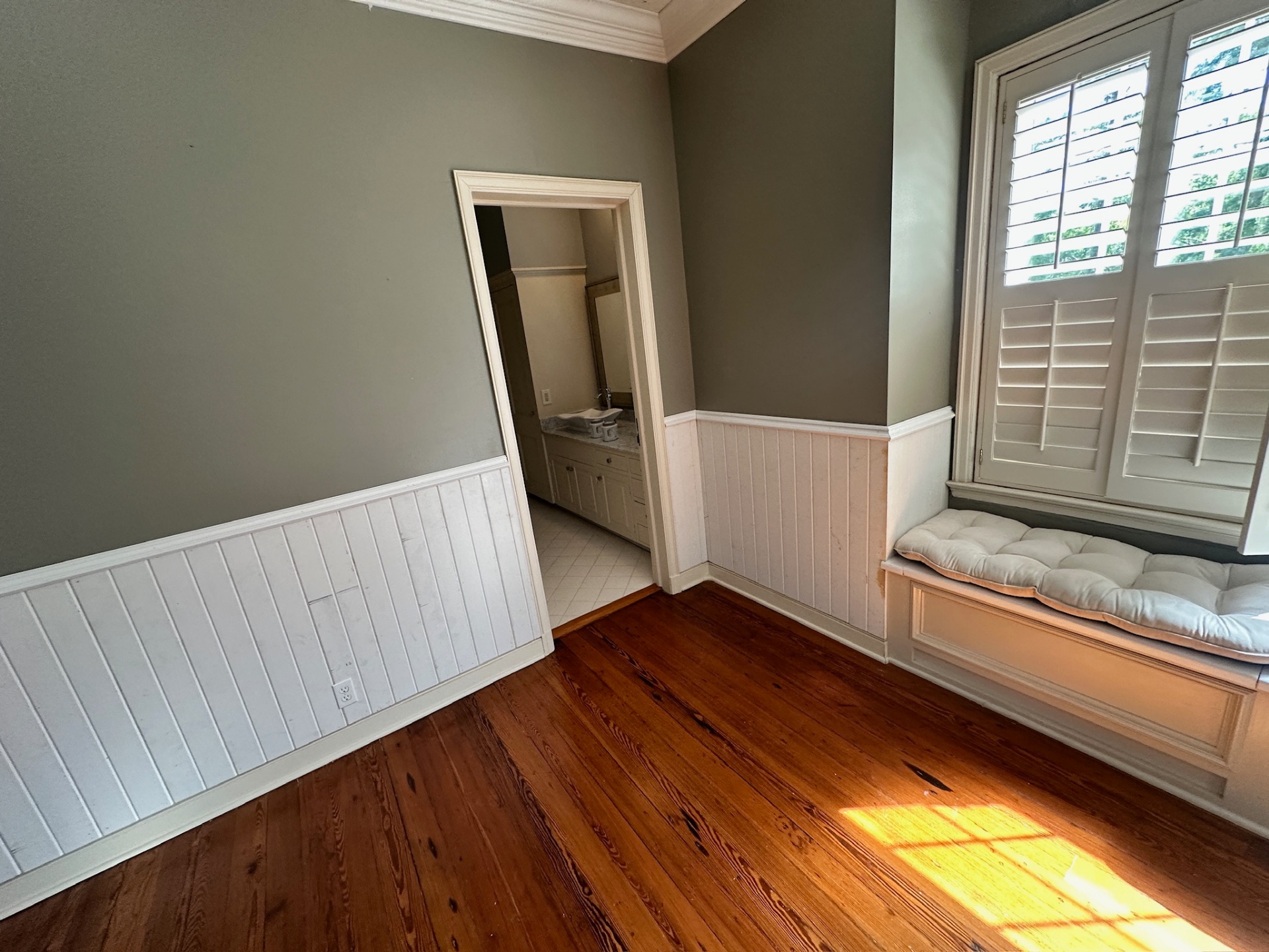 Finished bedroom nook in Covington LA showing custom white beadboard wainscoting scribed to reclaimed heart pine flooring next to an en-suite bathroom doorway and cushioned window seat