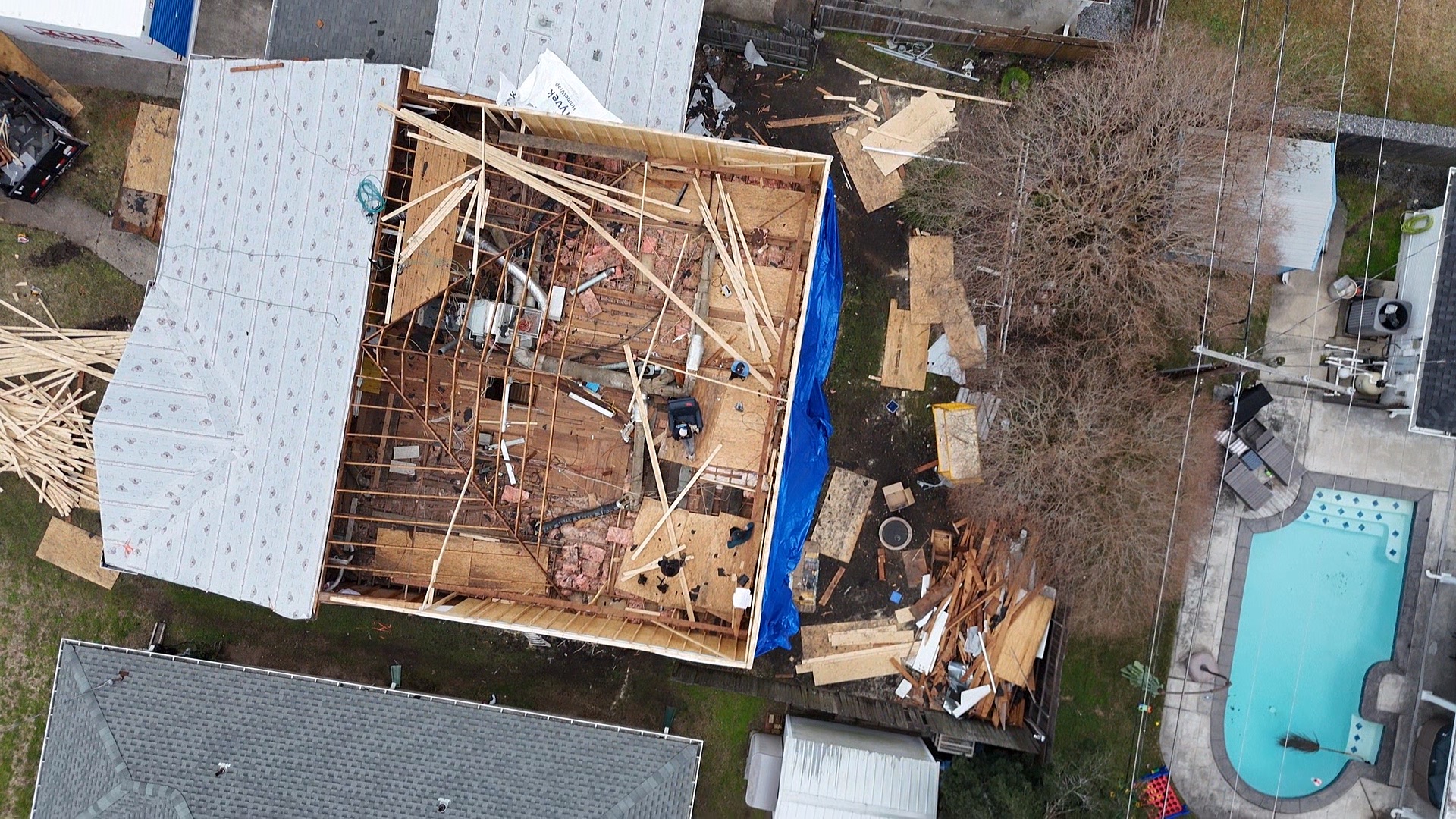 Aerial overhead drone view of Metairie home with roof removed showing original ceiling joists, blue tarp side protection and Tyvek-wrapped second-floor walls in progress
