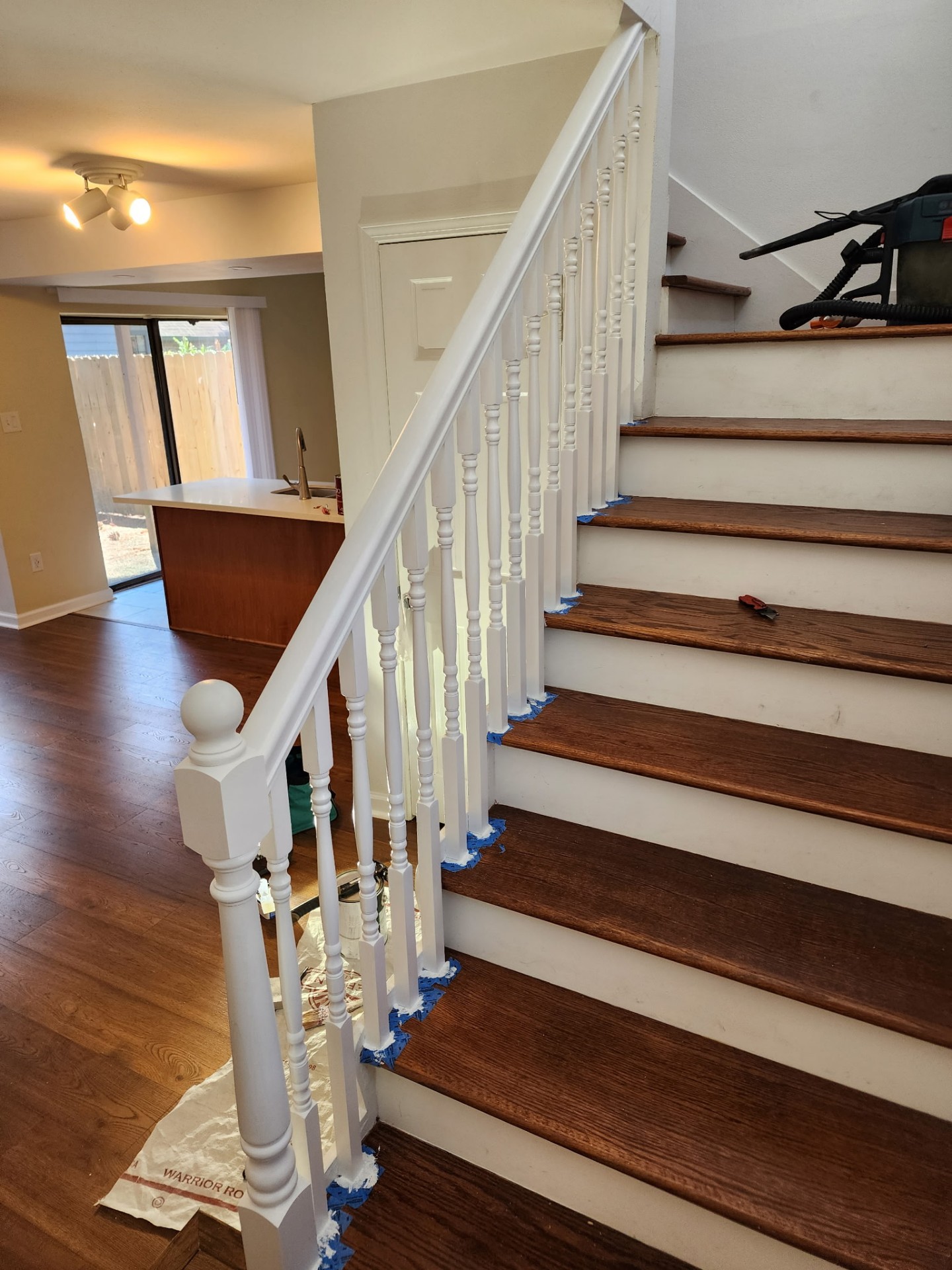 In-progress custom stair rail build in Kenner Louisiana showing installed newel post, turned balusters, and top mounted handrail with blue painter tape masking on the oak treads before final paint