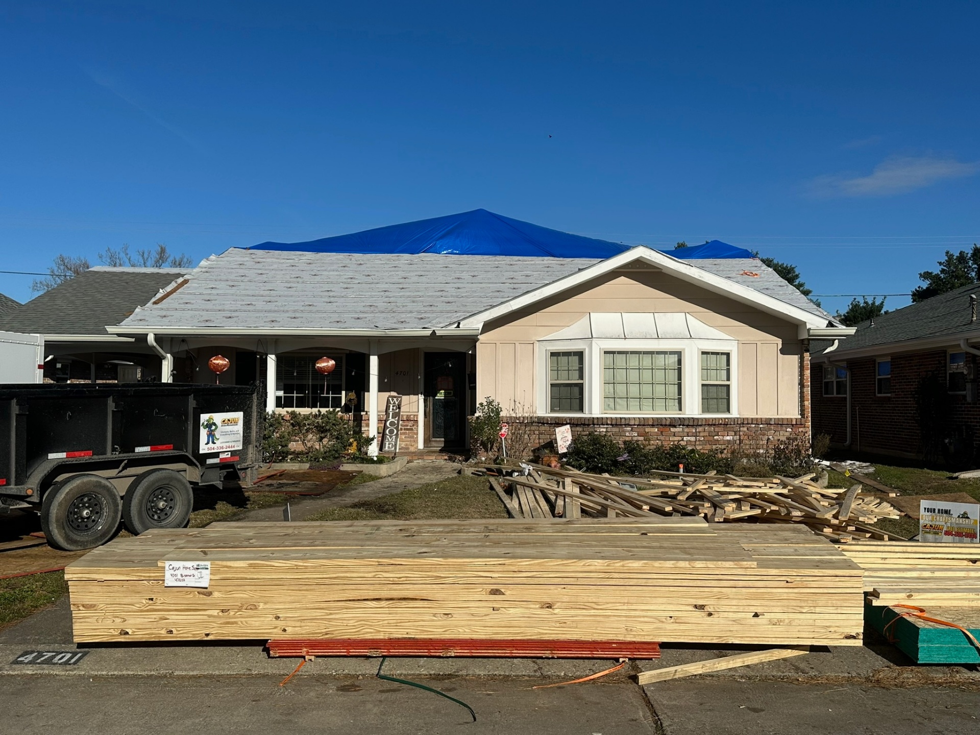 Front elevation of Metairie second-floor addition with synthetic underlayment dry-in installed and a blue tarp pulled over the peak, Cajun Home Services dump trailer at the curb and pressure-treated lumber staged in the driveway