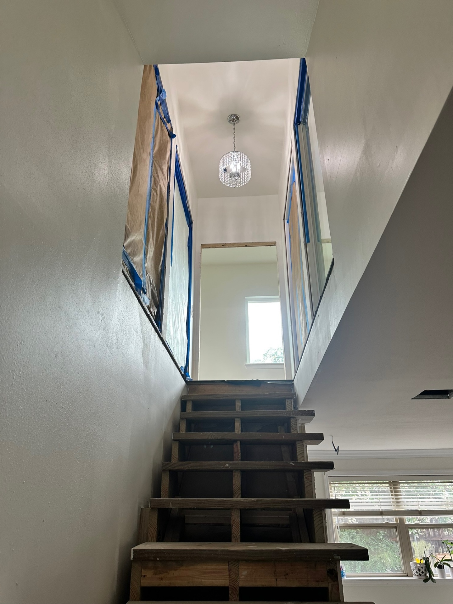Looking up the new open stair stringers from the first floor of the Metairie addition with raw treads in place, masked side wall, crystal chandelier in the upper landing and existing first-floor window with potted plant at right