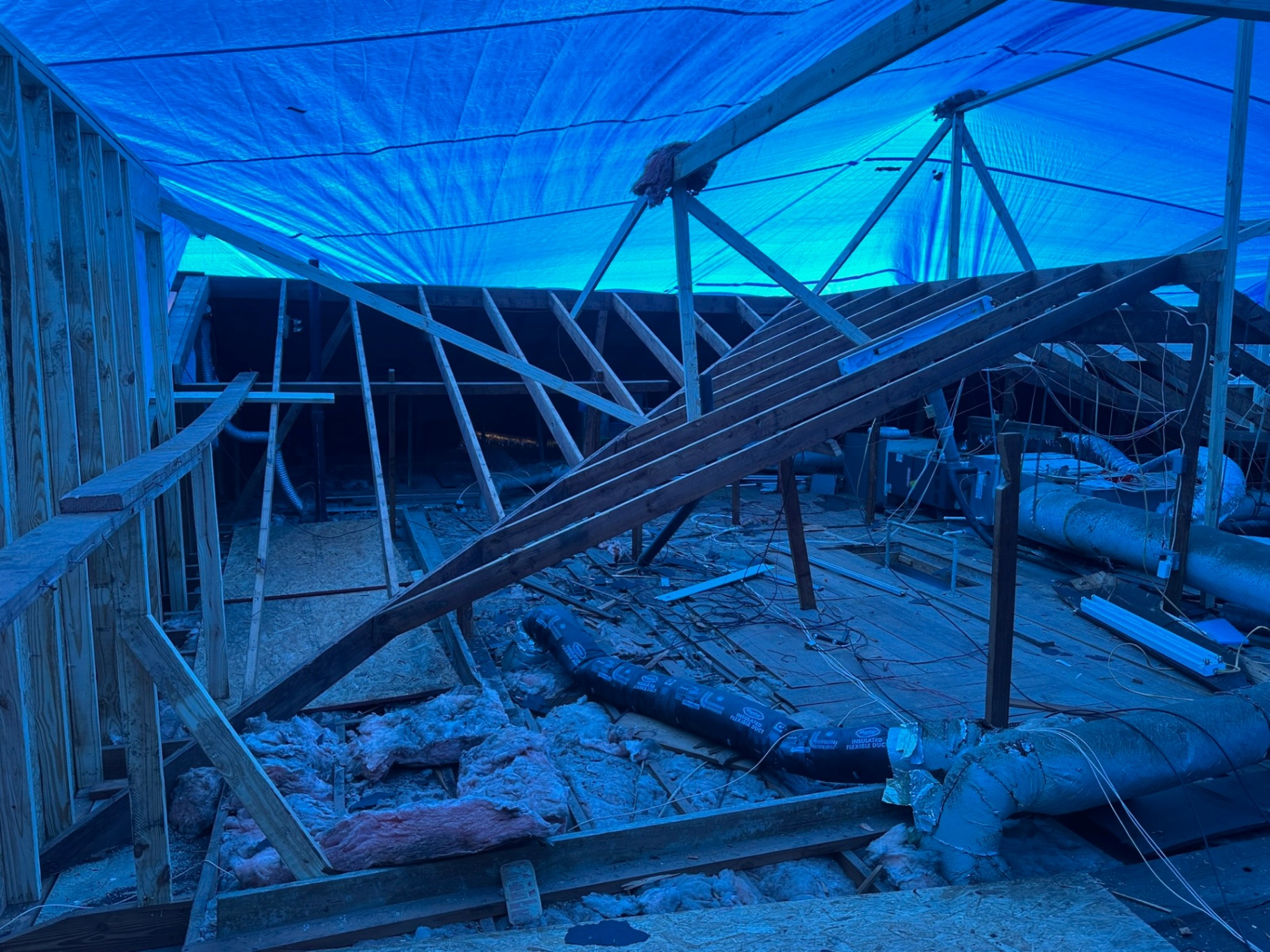 Inside Metairie attic during tear-off with old rafters partially collapsed onto floor joists, ductwork exposed and full blue tarp roof glowing overhead protecting the home
