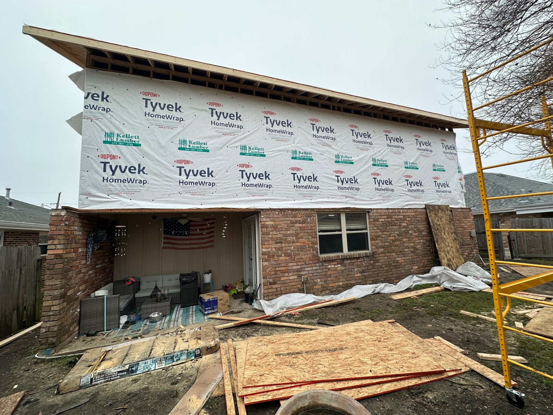 Wide rear elevation of Metairie second-floor addition fully wrapped in Tyvek with new roof framing visible above original brick first floor and scaffolding on site