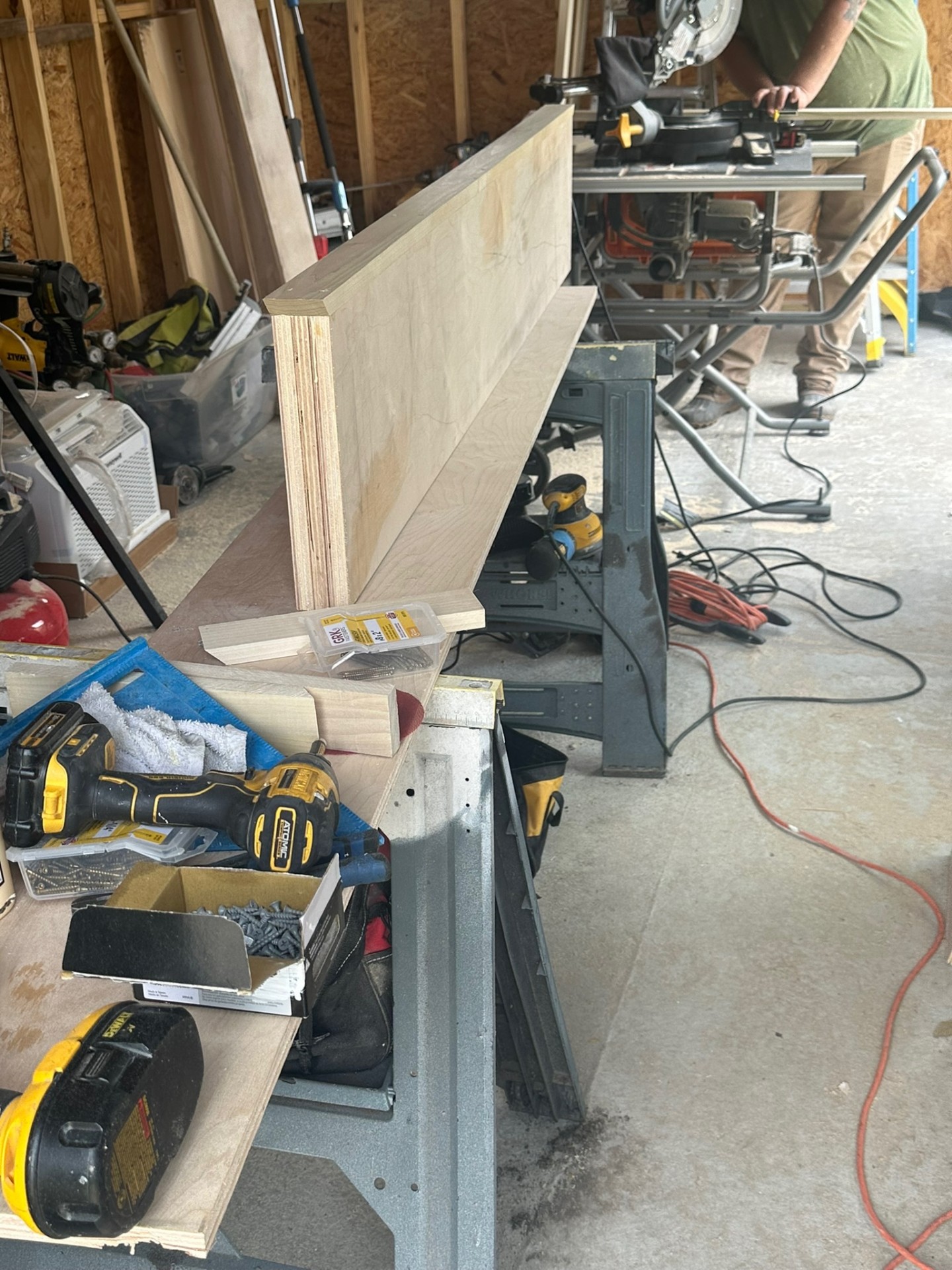 Cajun Home Services carpenter dry-fitting a solid maple floating shelf box on sawhorses in the shop while the miter saw is set up in the background for a custom shelf build in Meraux Louisiana