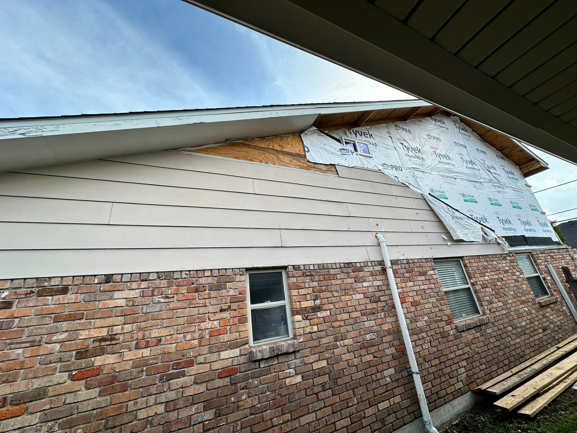 Side elevation of Metairie home addition mid-siding install showing new Hardie lap siding painted tan on the second floor, original brick first floor below and remaining Tyvek and Kellett Lumber sheathing exposed at the gable end