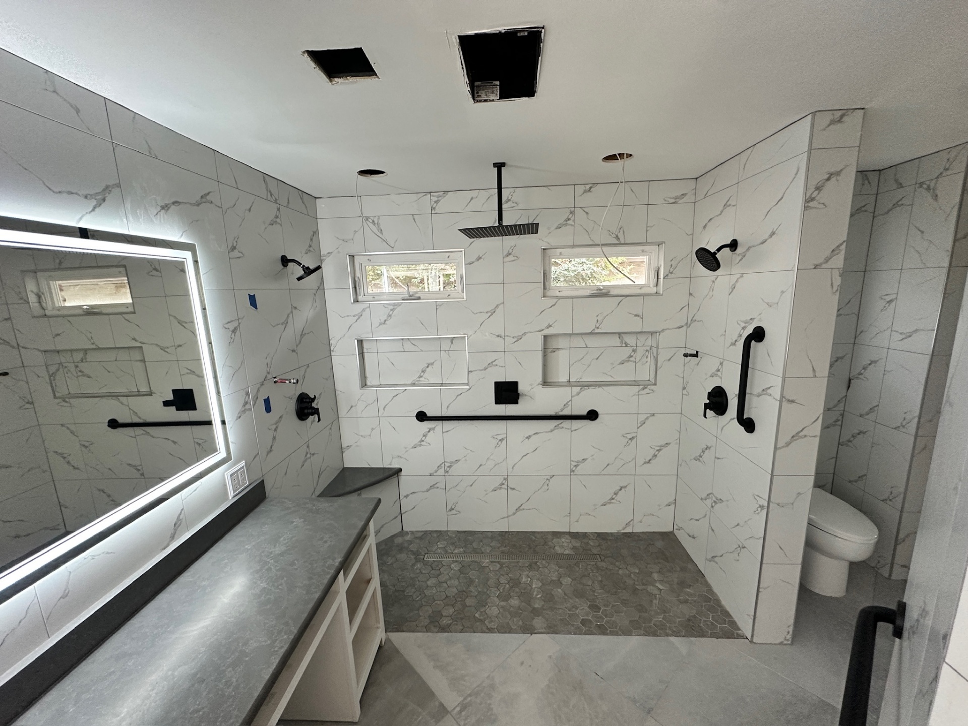 Walk-in shower with dual heads, dual niches, black grab bars, hex floor, and LED-mirror vanity installed before glass doors in Metairie master bath