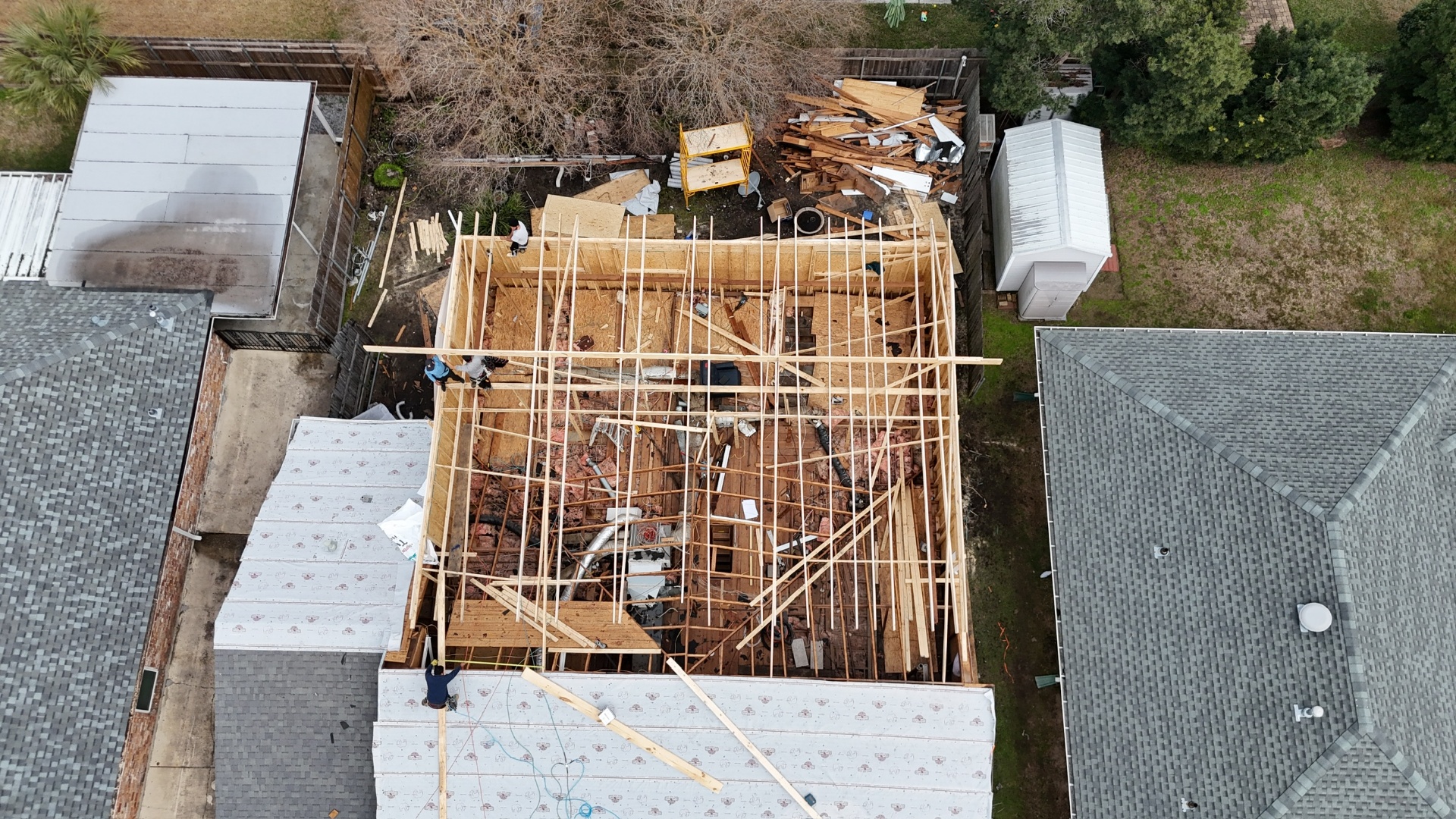 Overhead aerial view straight down on Metairie second-floor addition with new roof rafters being set across Tyvek-wrapped walls and lumber piled in the yard