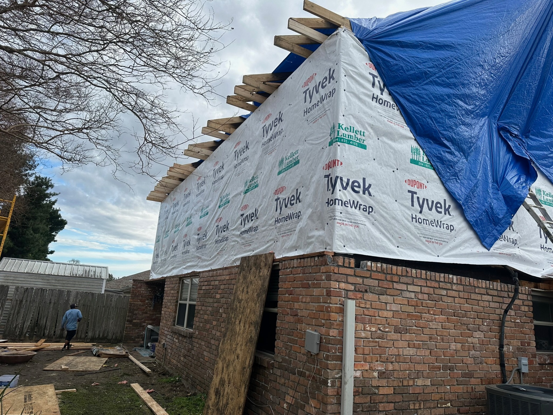 Side gable end of Metairie second-floor addition wrapped in Tyvek with exposed rafter tails and blue tarp draped over the ridge before roofing