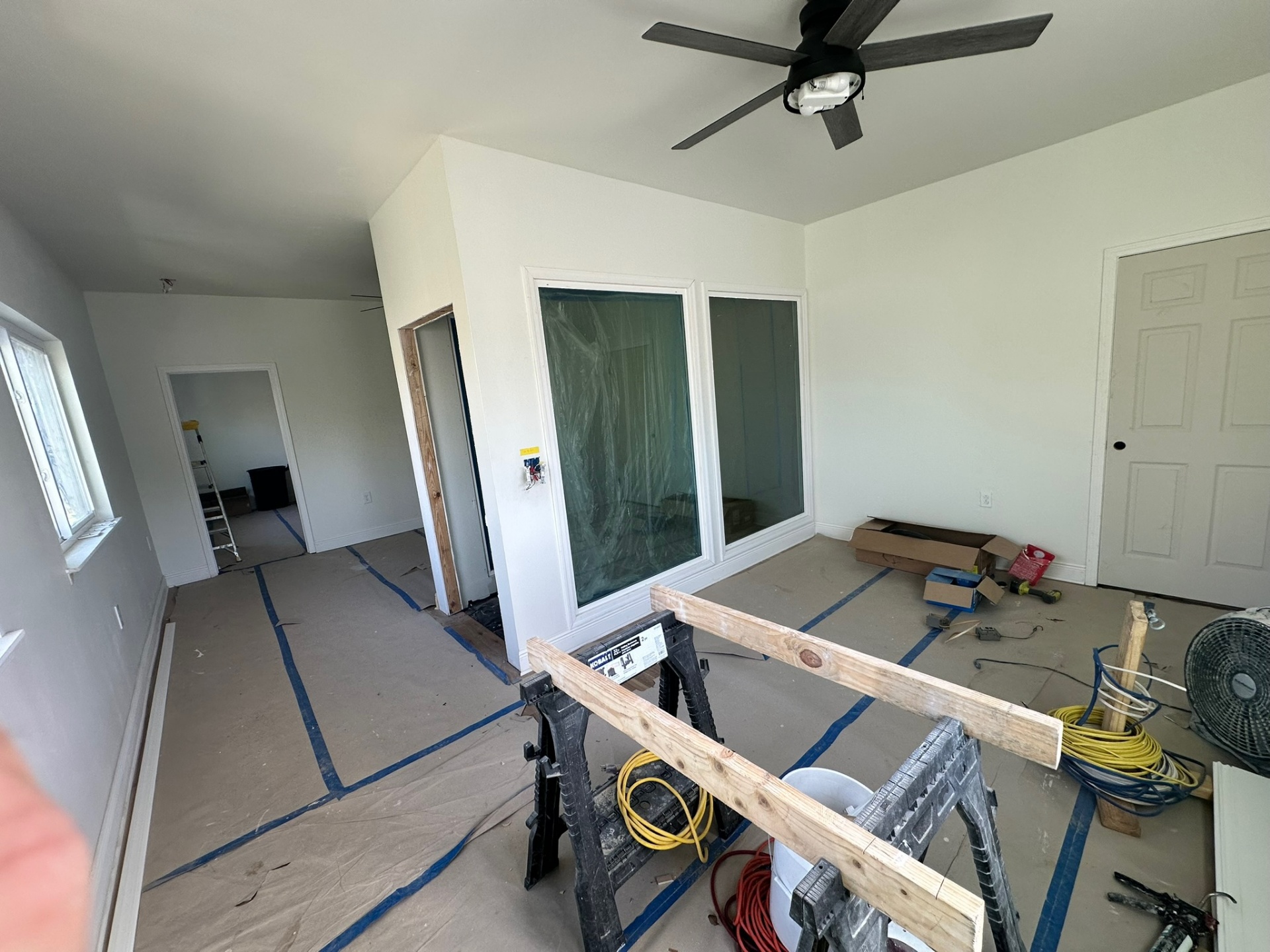 Second-floor bedroom near completion with new interior sliding glass door, dark ceiling fan, framed pocket door rough opening and finish carpentry materials on sawhorses