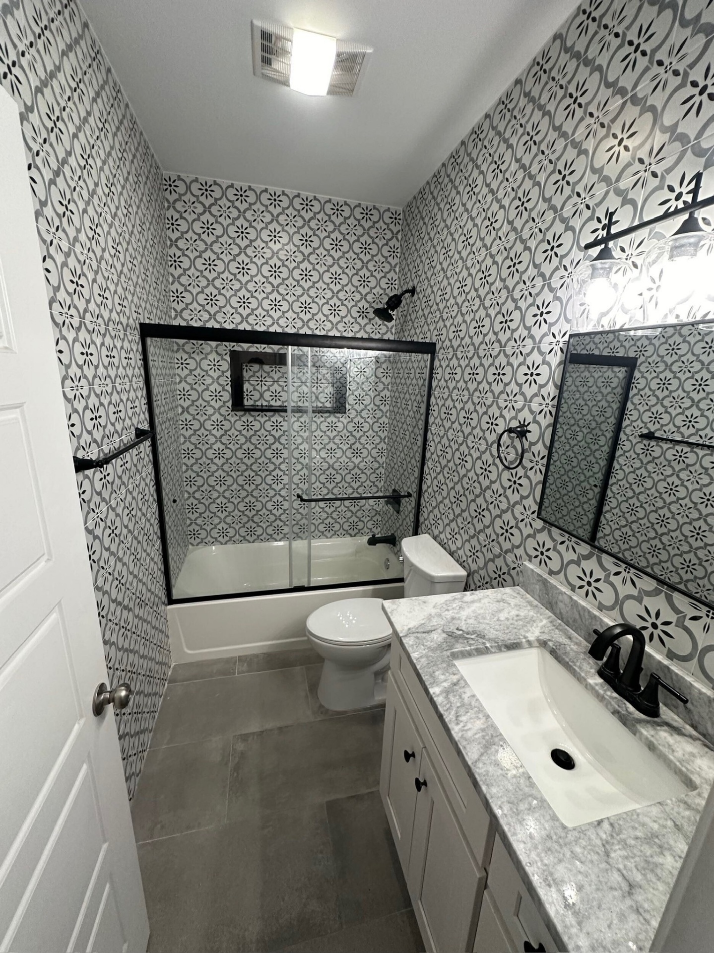 Finished hall bathroom in Meraux Louisiana with floor to ceiling patterned black and white porcelain tile on the tub surround, gray floor tile, framed glass sliding tub door, marble top vanity with black faucet, and black framed mirror