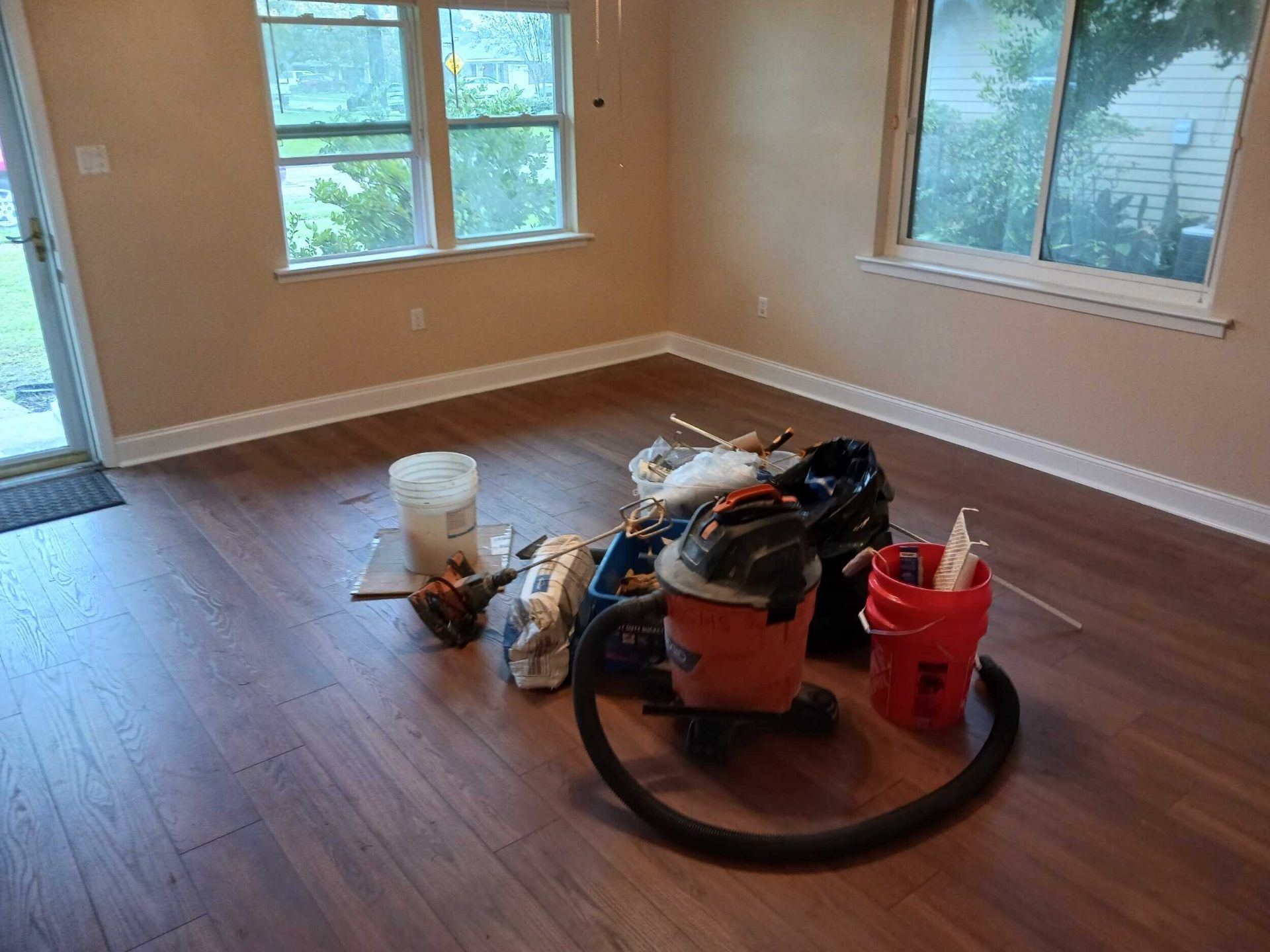 Wide view of the finished rebuilt Gretna living room with new windows, fresh paint, new laminate flooring, and the crew's tools staged for punch-list walkthrough