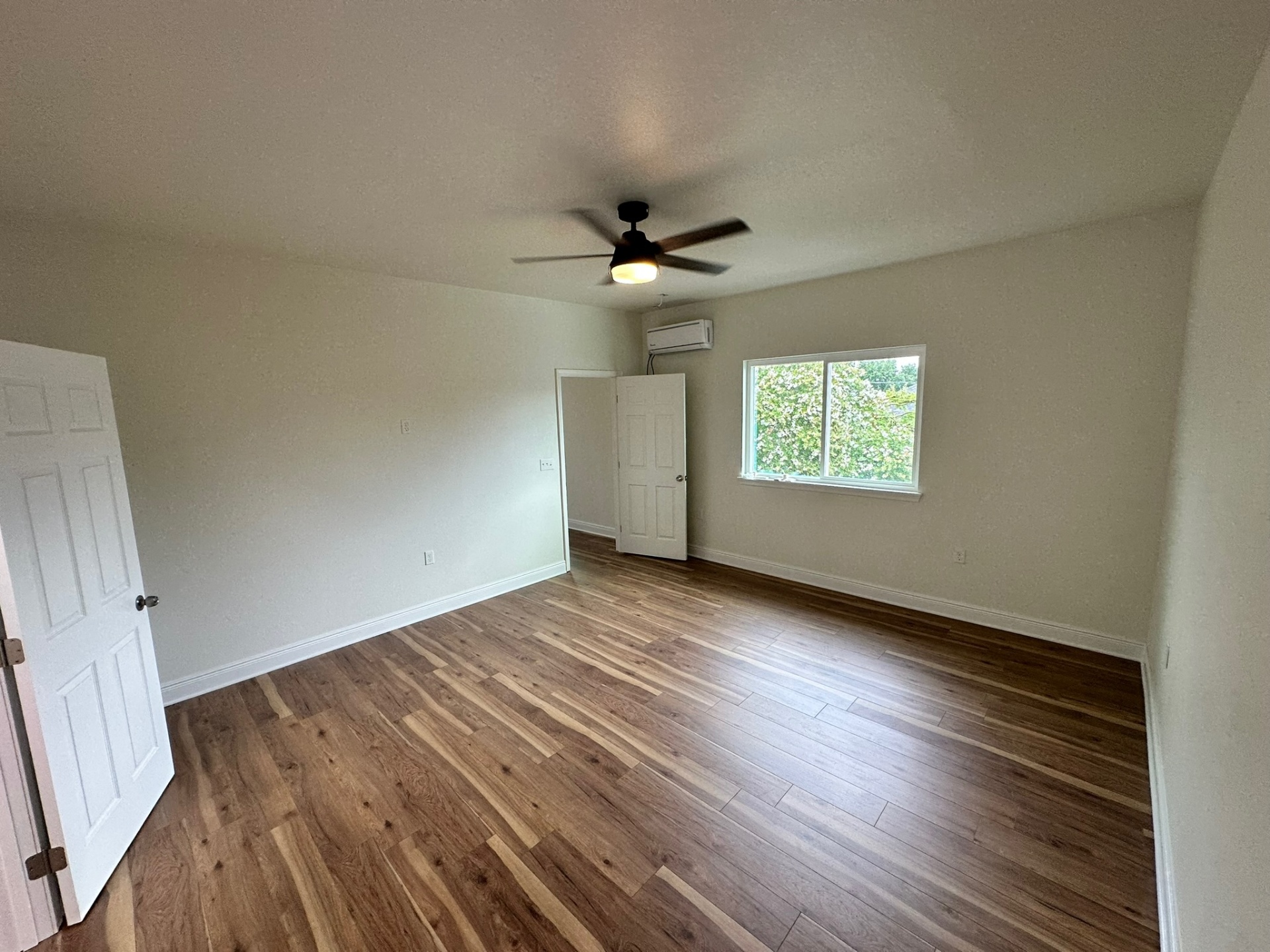 Finished new second-floor primary bedroom in the Metairie addition with light walls, wide-plank LVP flooring, ductless mini-split, large slider window over landscaping, ceiling fan with light and door open to the en-suite hallway