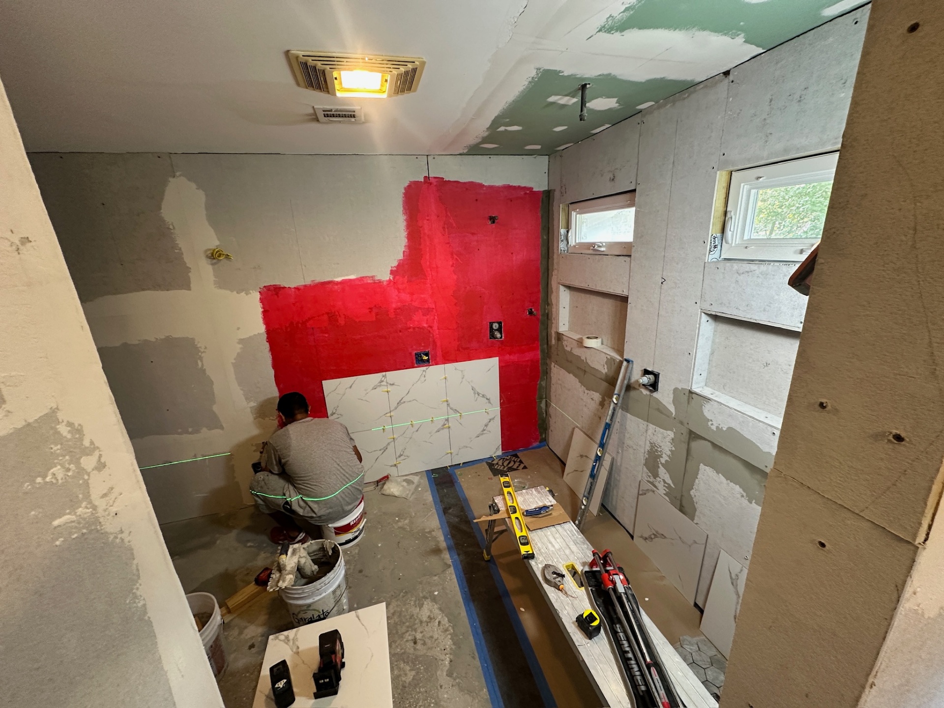 RedGard waterproofing rolled onto shower walls with first row of marble-look tile going up and laser level in Metairie master bath