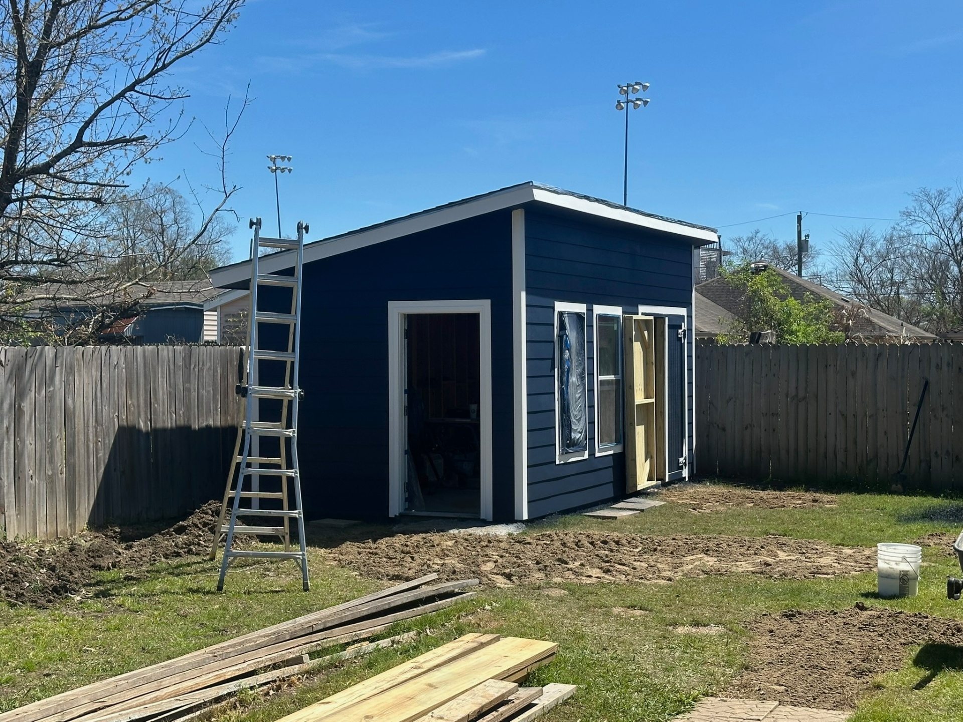 Wide view of nearly completed Metairie shed with extension ladder, lumber stack, and graded backyard ready for sod