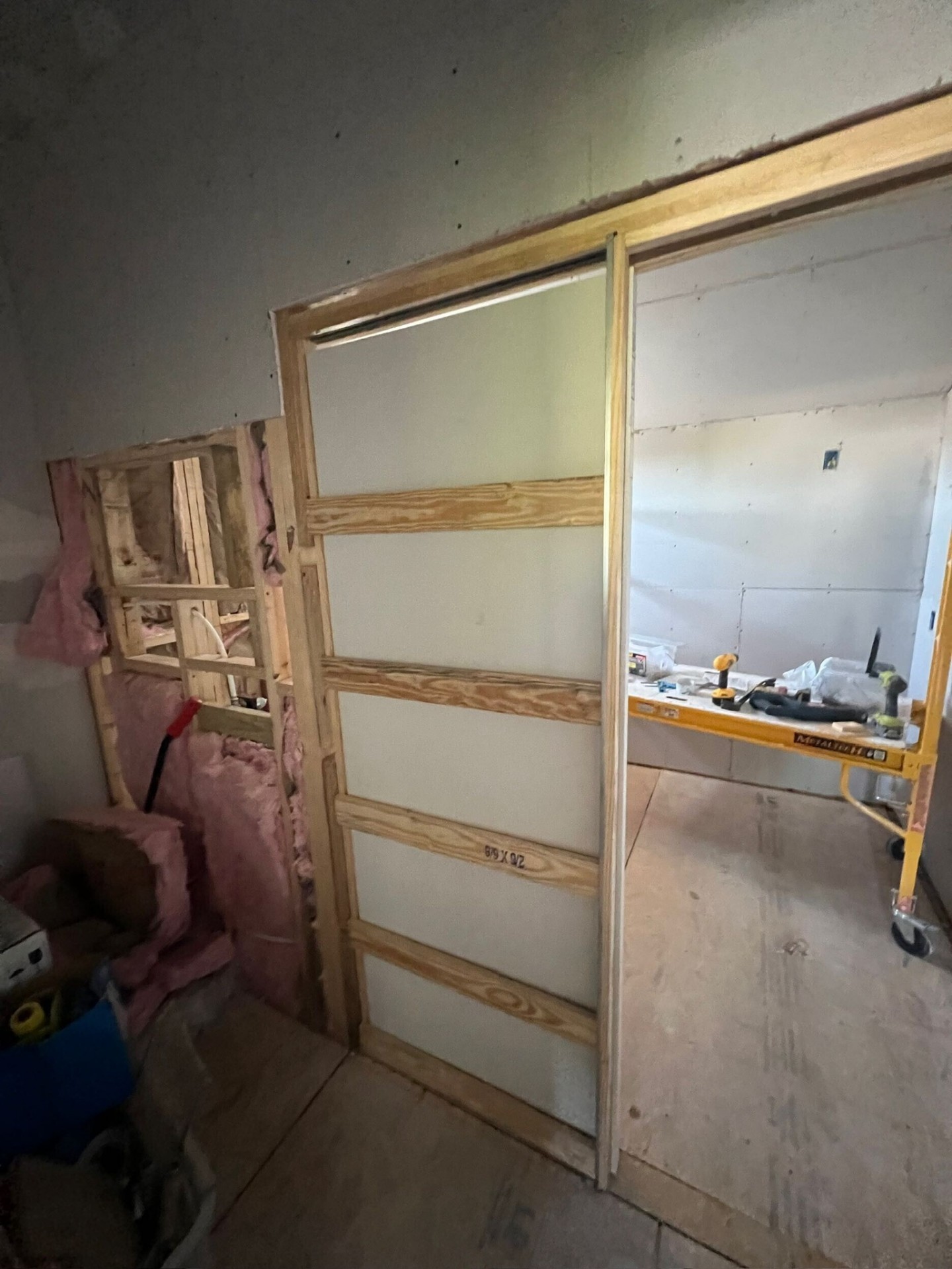 Second-floor pocket door rough frame standing in a new partition wall on the Metairie addition with pink fiberglass insulation in the adjacent stud bay and a DeWalt drill on a yellow work table in the next room